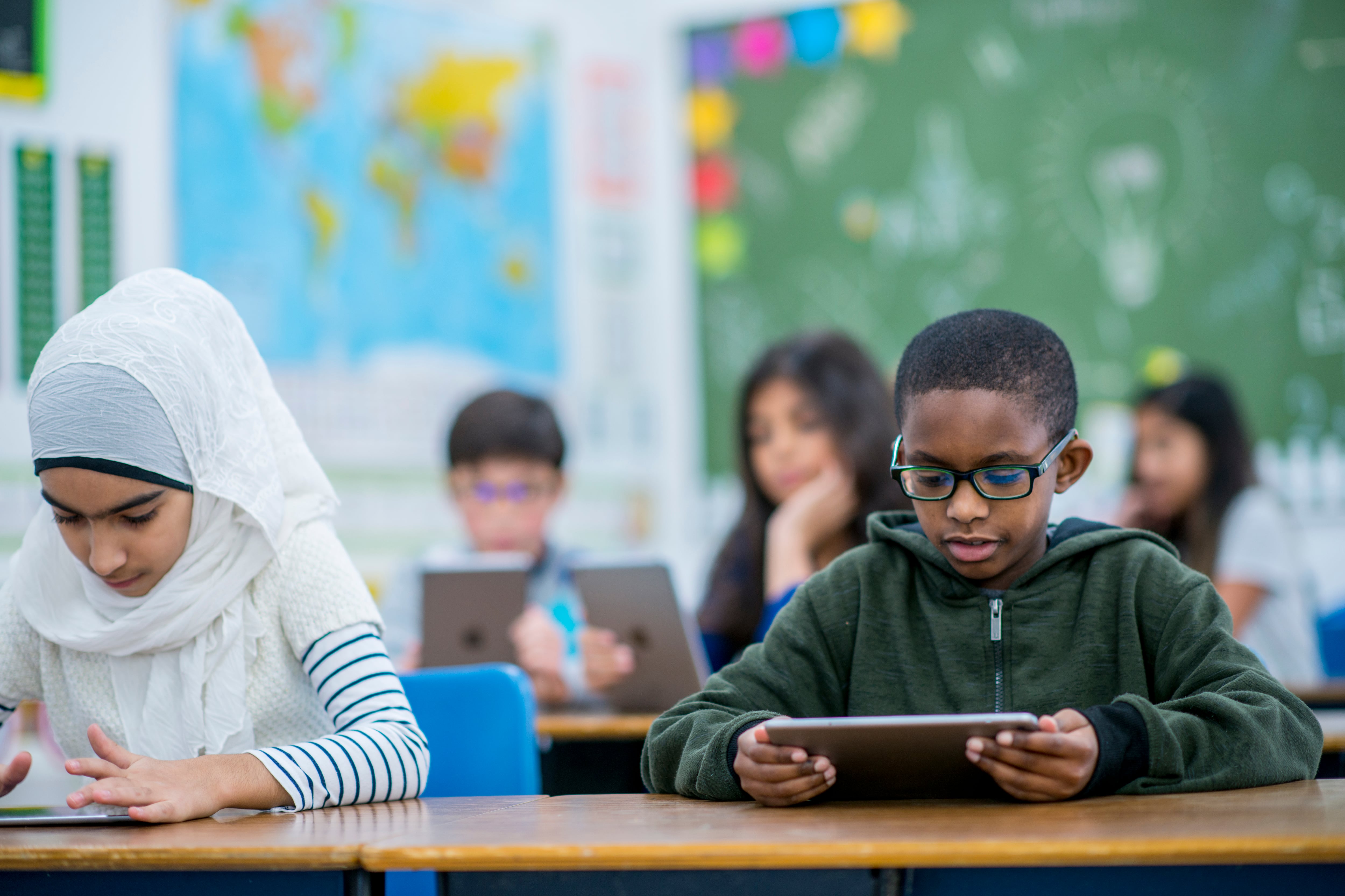 A young girl wearing a white hijab sits at a desk working on a tablet computer. She sits next to a young boy wearing a green sweatshirt with black glasses who is also working on a tablet.