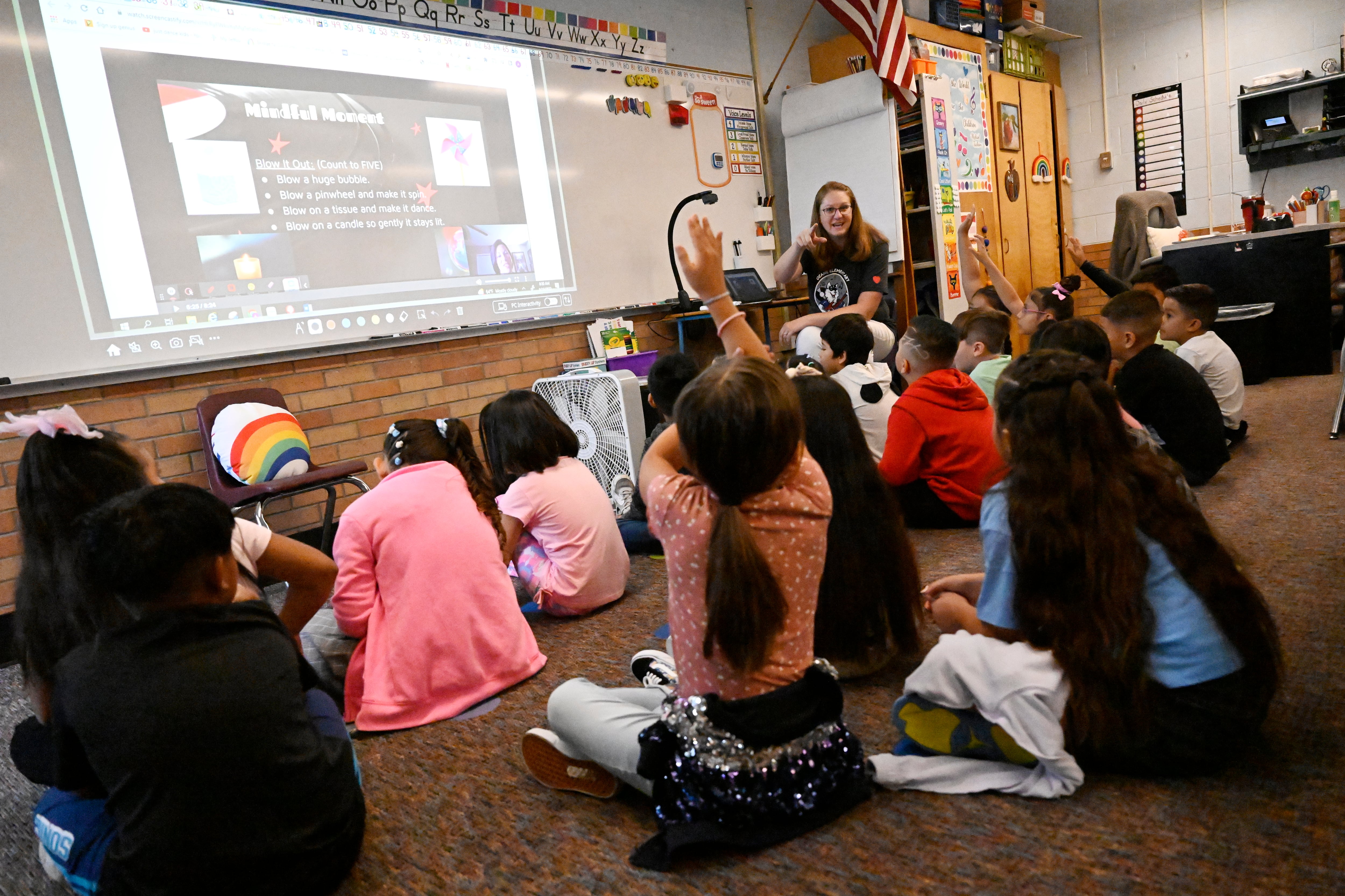 A group of students sits on the floor looking at their teacher. There’s a presentation projected on the wall. One girl has her hand raised as the teacher speaks to the class.