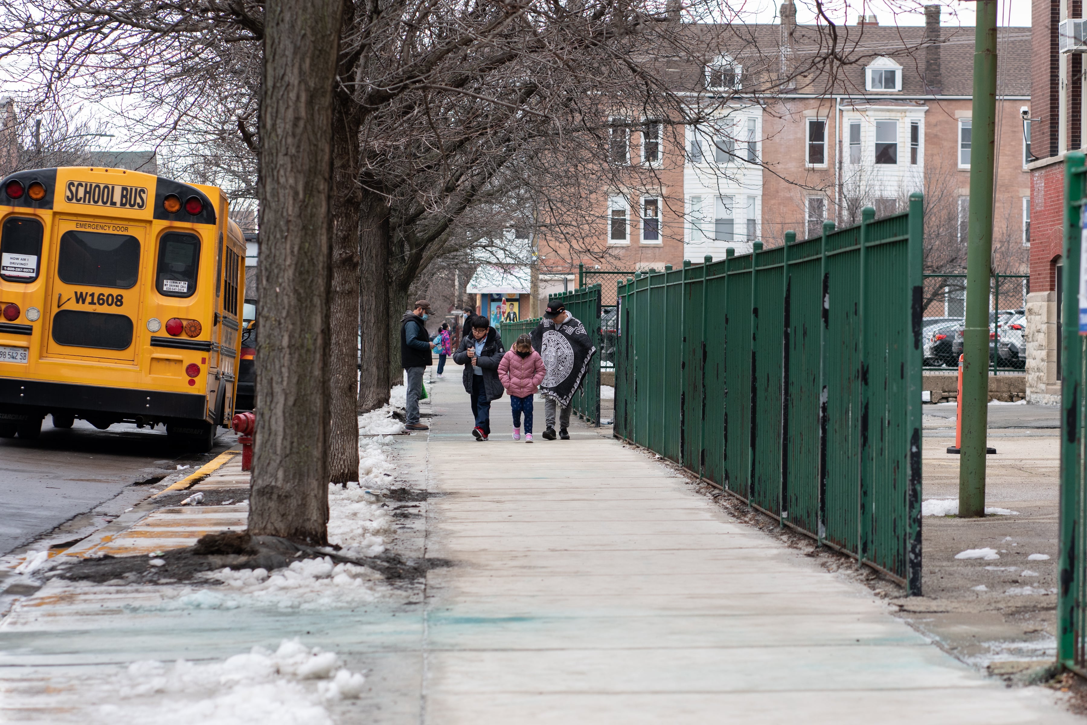 A man and two children walk down a sidewalk by a yellow school bus.