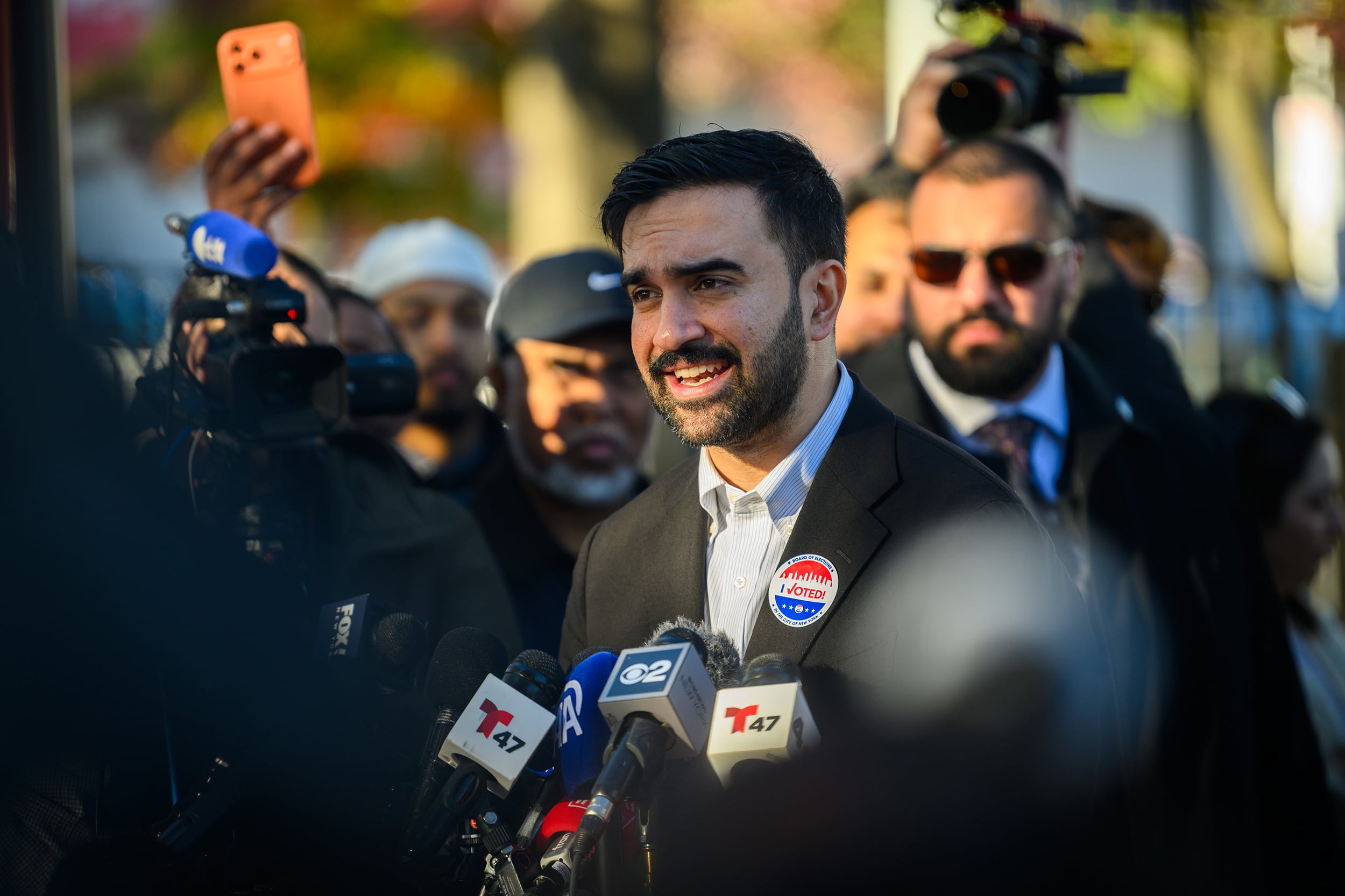 A photograph of a man in a suit speaking in front of microphones in a large group of people outside.
