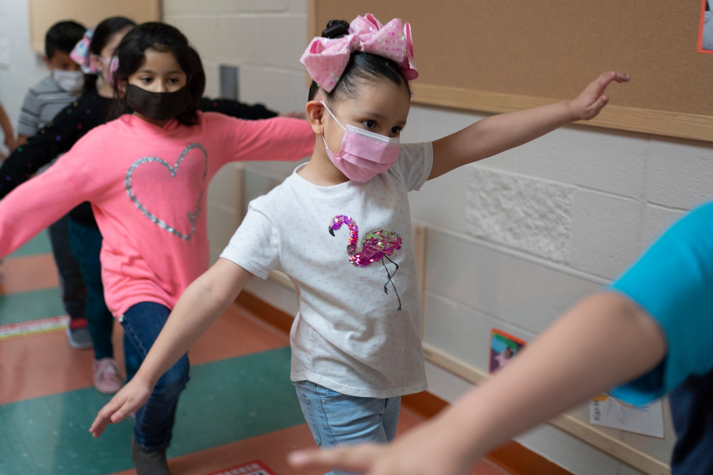 A child wearing a blue shirt is in front of a girl wearing a white flamingo shirt, a pink bow, and a pink surgical mask, arms outstretched in a class hallway. The girl behind her is wearing a pink sweater with a silver heart on it.