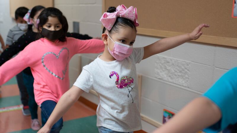 A child wearing a blue shirt is in front of a girl wearing a white flamingo shirt, a pink bow, and a pink surgical mask, arms outstretched in a class hallway. The girl behind her is wearing a pink sweater with a silver heart on it.