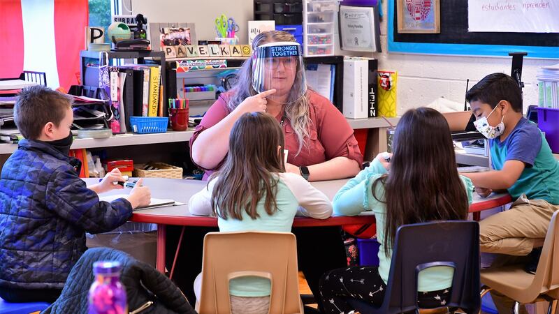 A teacher sits at a table facing four students. The teacher wears a clear plastic face shield and points to her chin.