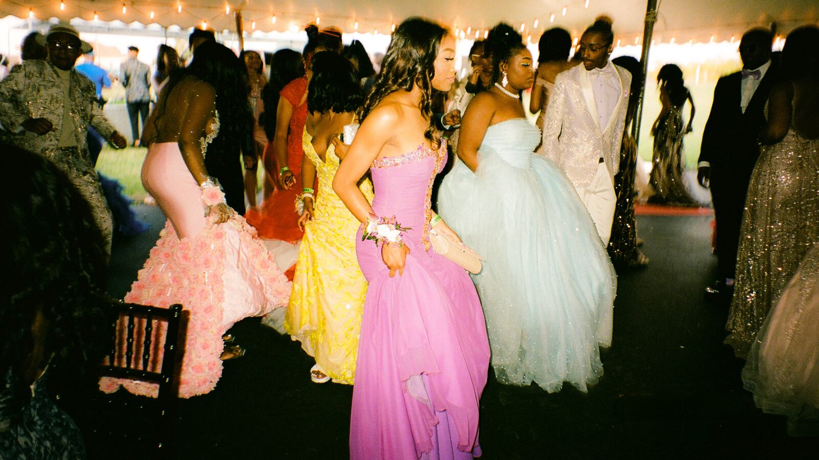 Teenagers dance together under a party tent, wearing a wide array of colorful dresses and suits as string lights illuminate the inside of the tent.