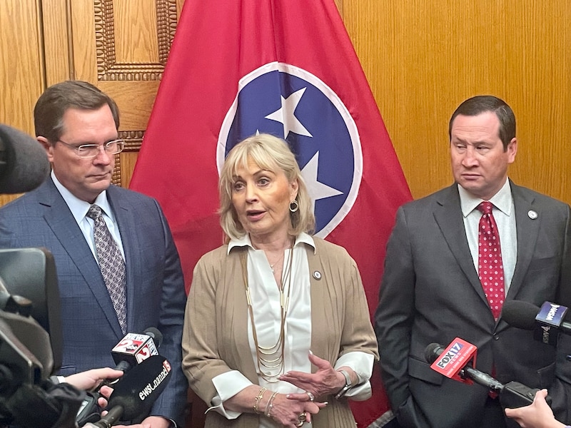 Three lawmakers address media. A woman in the center, speaking, wears a white collared shirt and tan cardigan.