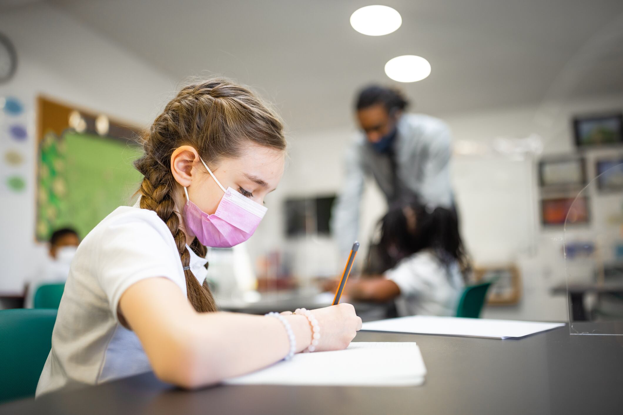 A girl in a mask writes on a white piece of paper with a pencil.