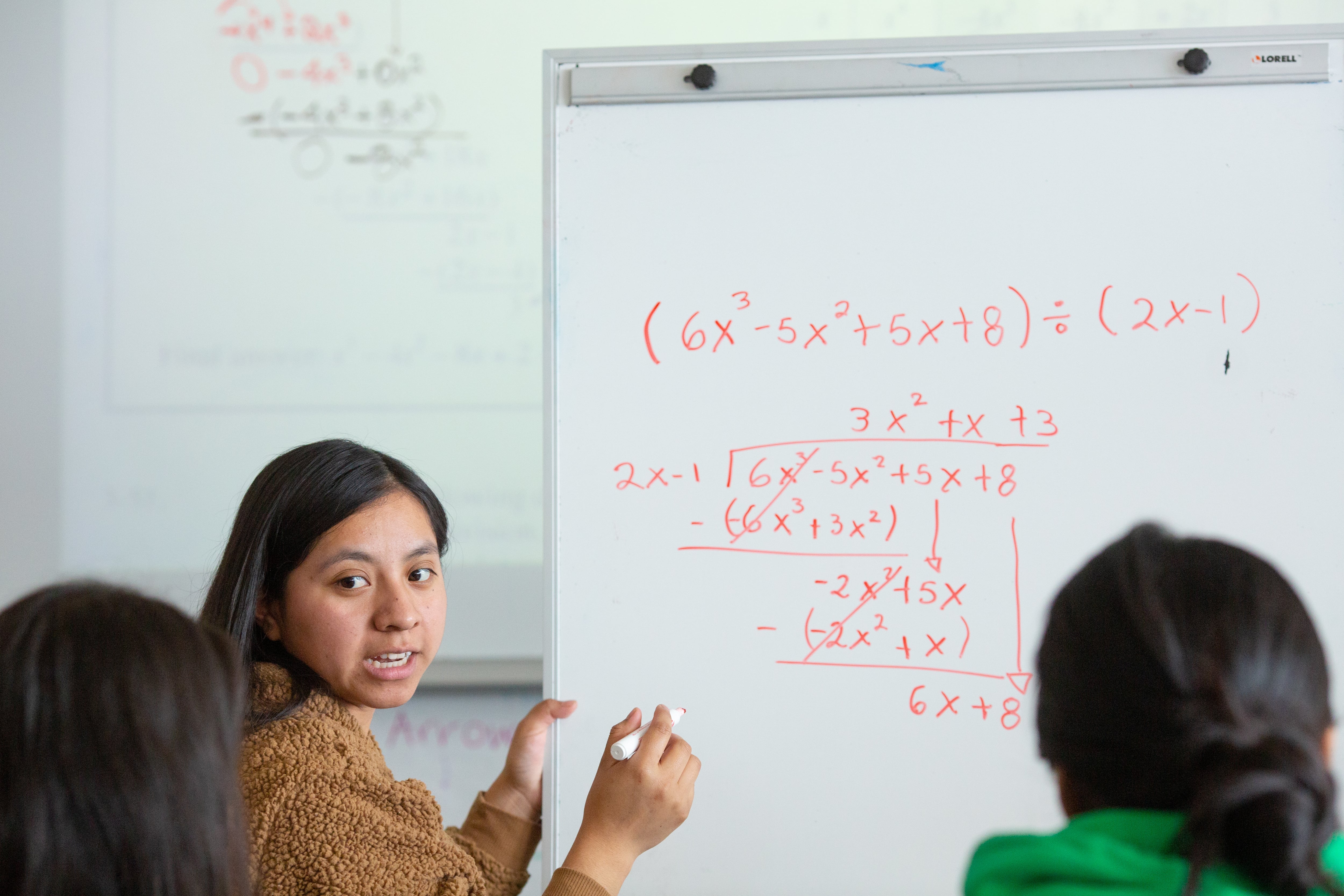 A girl with long brown hair stands at a whiteboard solving an algebra equation. The equation is written in red marker on the board.