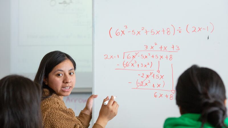 A girl with long brown hair stands at a whiteboard solving an algebra equation. The equation is written in red marker on the board.