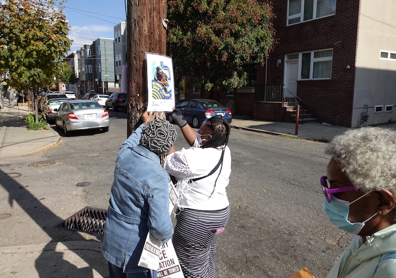Two people attached a poster to a telephone pole outside while a person stands in the foreground.