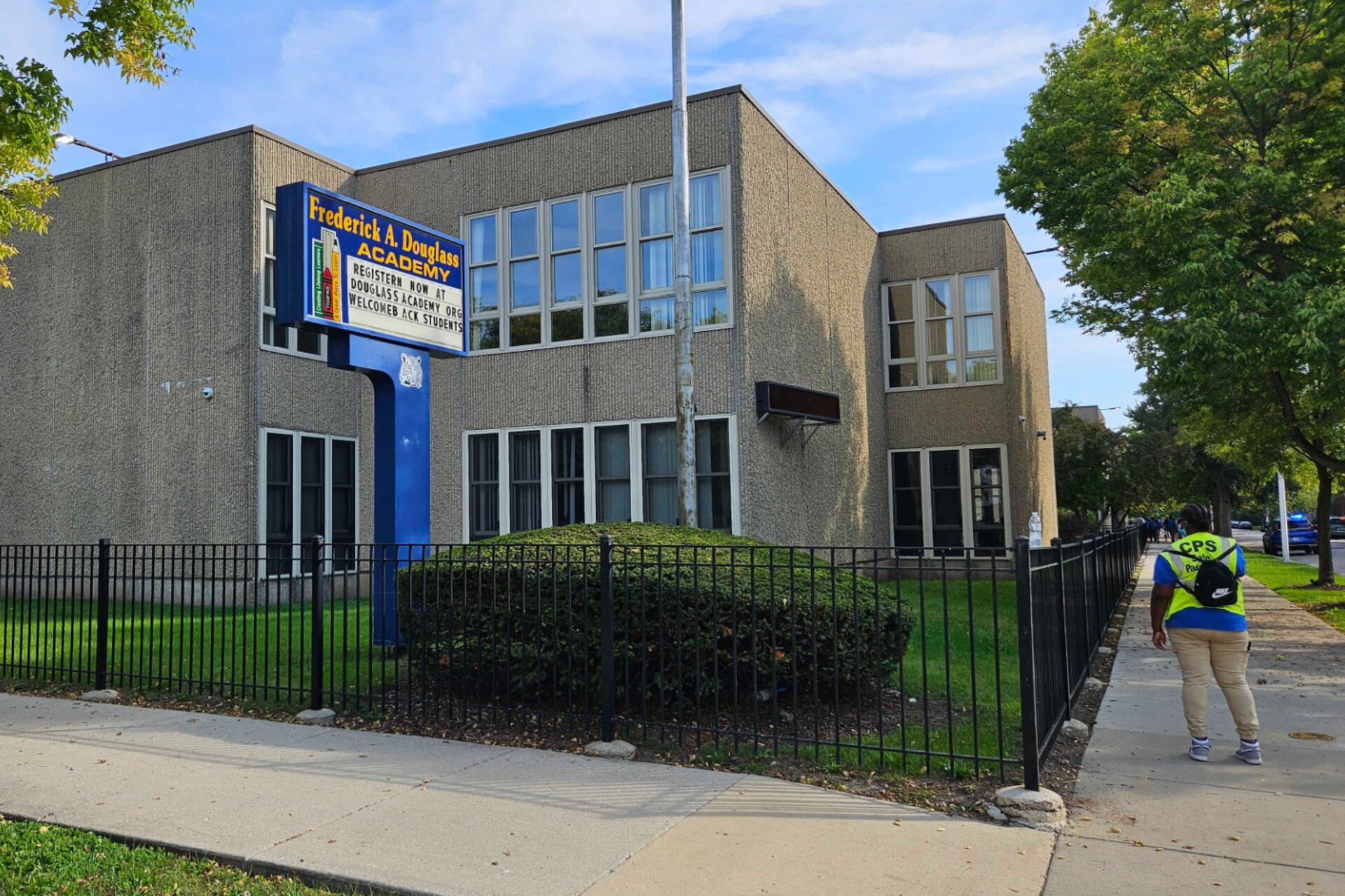 The outside of a school with the person to the right walking down the side walk. There is a large sign in the foreground and blue sky in the back ground.