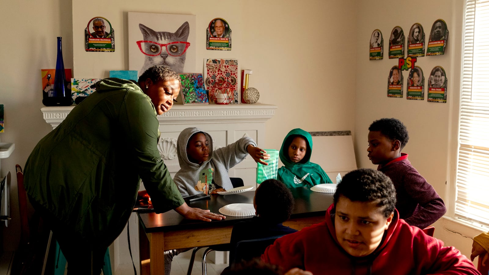 An adult woman wearing a green shirt stands next to two tables full of five young children sitting down in a house with nice light shining through a window.