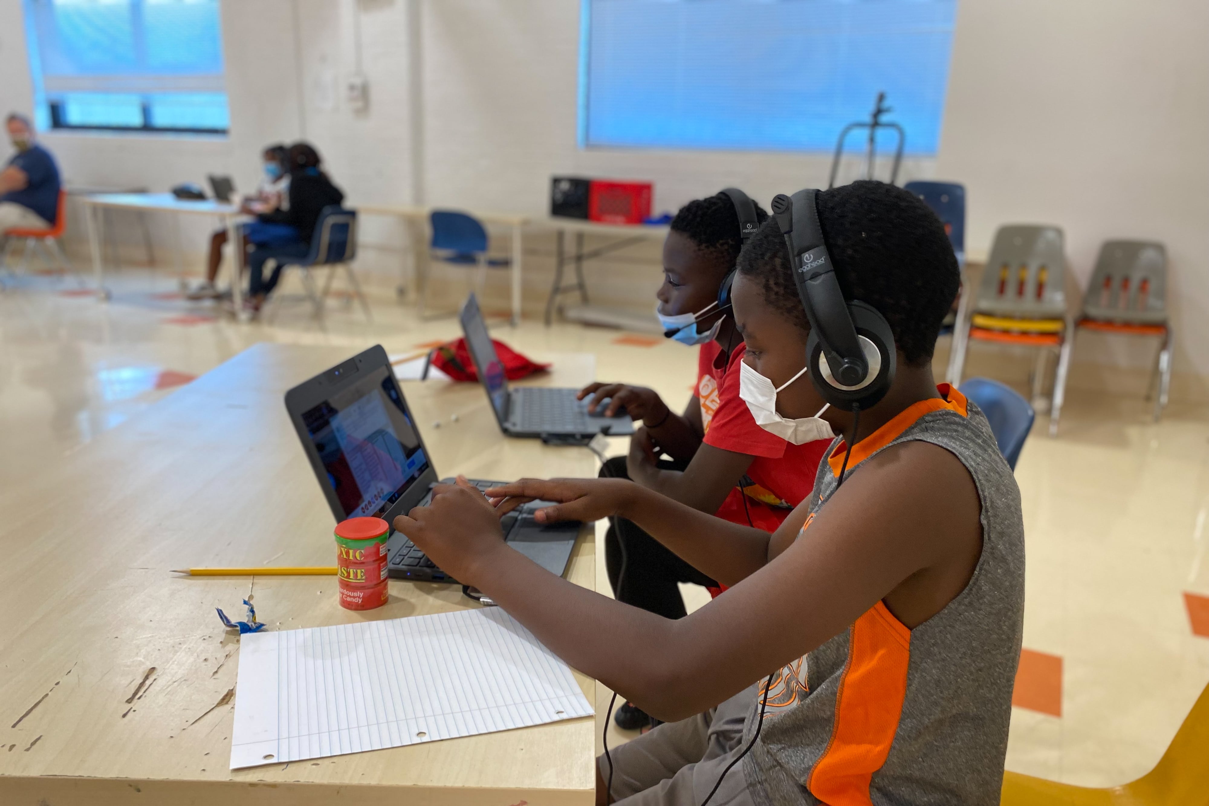 Two male students with masks on are sitting down in a large room typing on laptop computers.