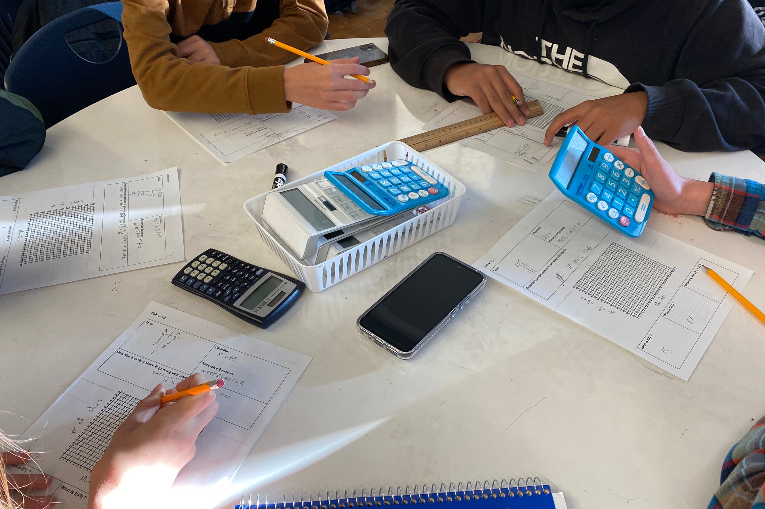 Four students sit at a table working on classwork. The table is white with papers and blue calculators.