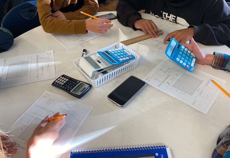 Four students sit at a table working on classwork. The table is white with papers and blue calculators.