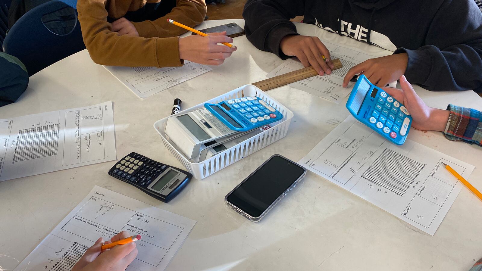 Four students sit at a table working on classwork. The table is white with papers and blue calculators.