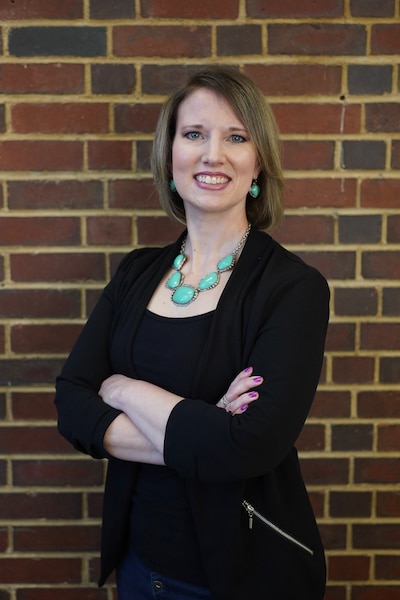 A photograph of a white woman with short blonde hair posing for a photograph in front of a brick wall.