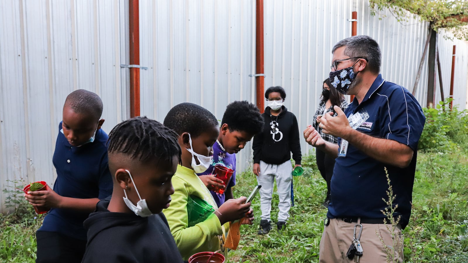 A teacher works in an outdoor space with several students, who are holding discovered seeds in plastic cups.