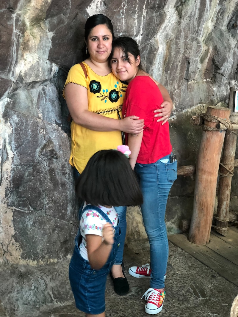 A mother wearing a yellow shirt stands near a rock formation and hugs her teenage daughter, while her younger daughter spins in front of them. The older daughter is wearing a red T-shirt and jeans.