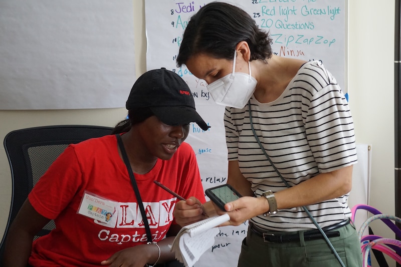 A photograph of a white woman wearing a mask and a white shirt with black stripes interviews a Black teen wearing a red shirt and a black baseball cap in a room.