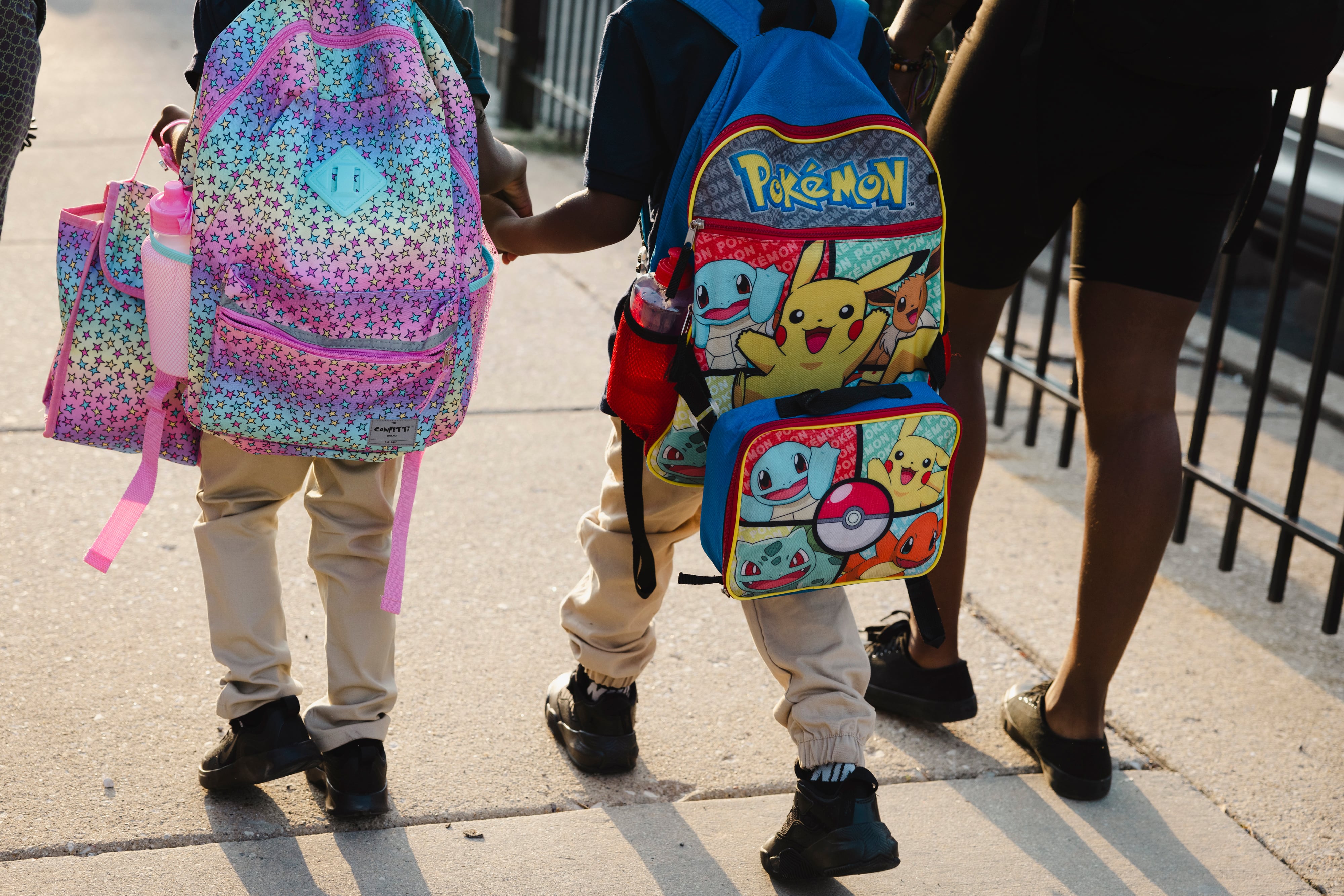 A rearview of two young students carrying colorful backpacks outside.