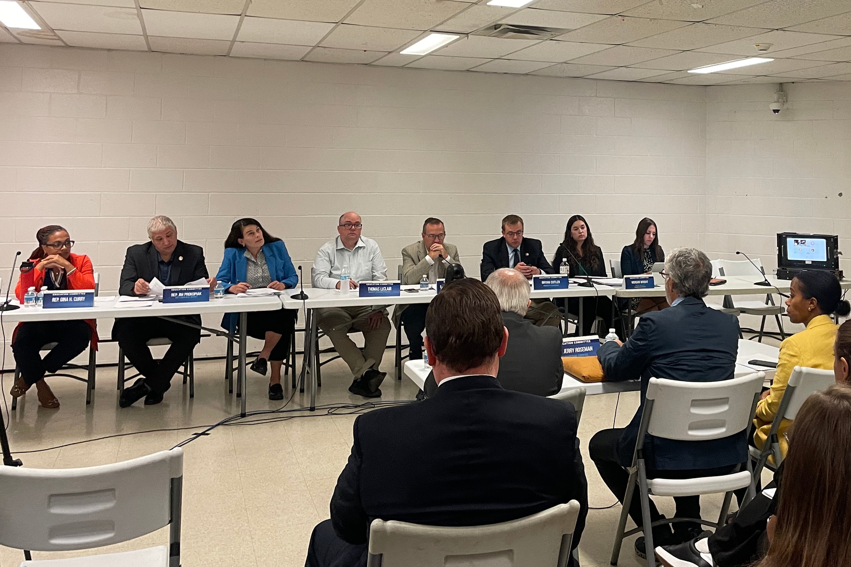 A photograph of a group of adults in business clothes sitting in a row in front of a seated audience in a community center room.