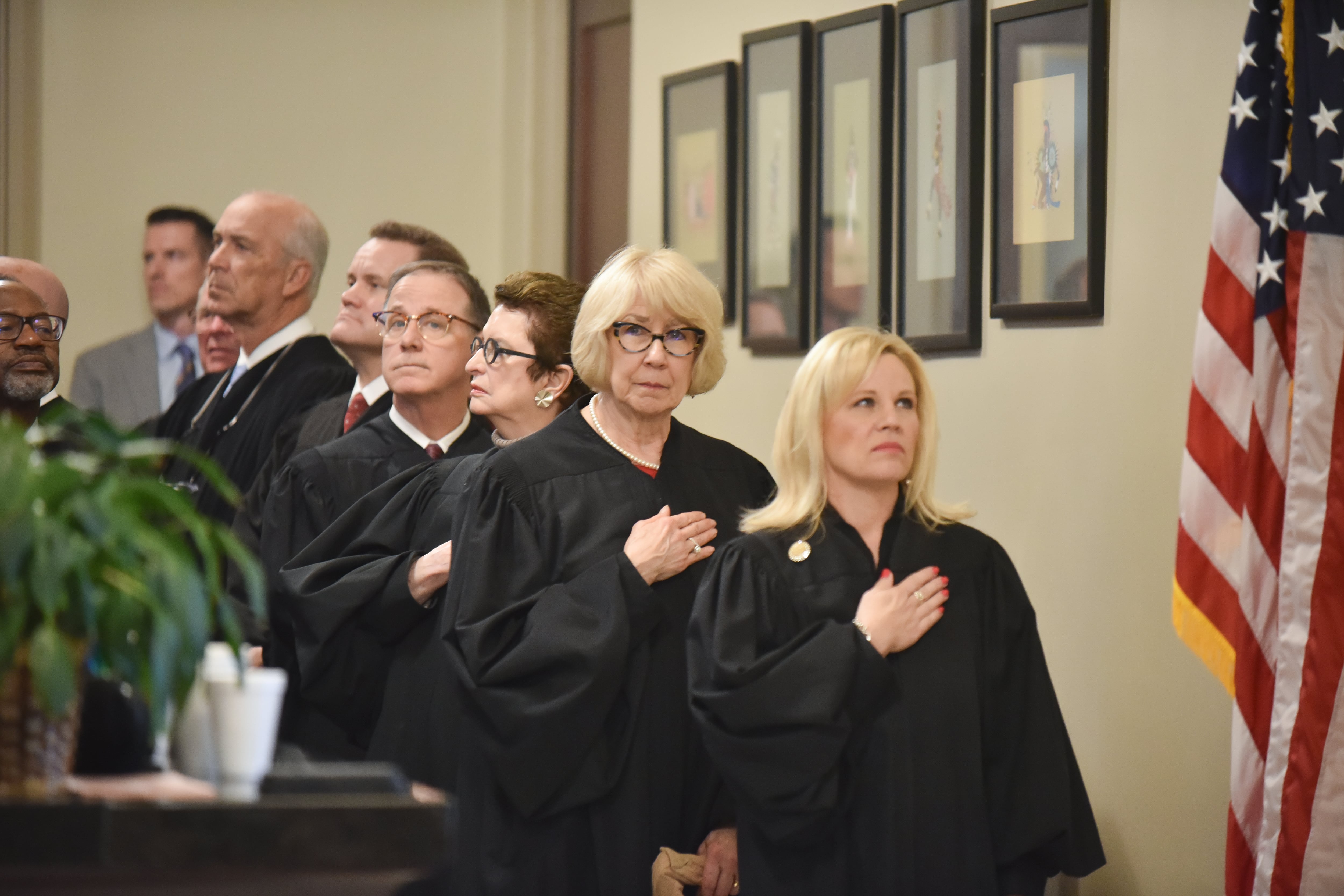 Oklahoma Supreme Court justices in black judicial robes place their hands over their hearts. An American flag is visible on the right side of the image.