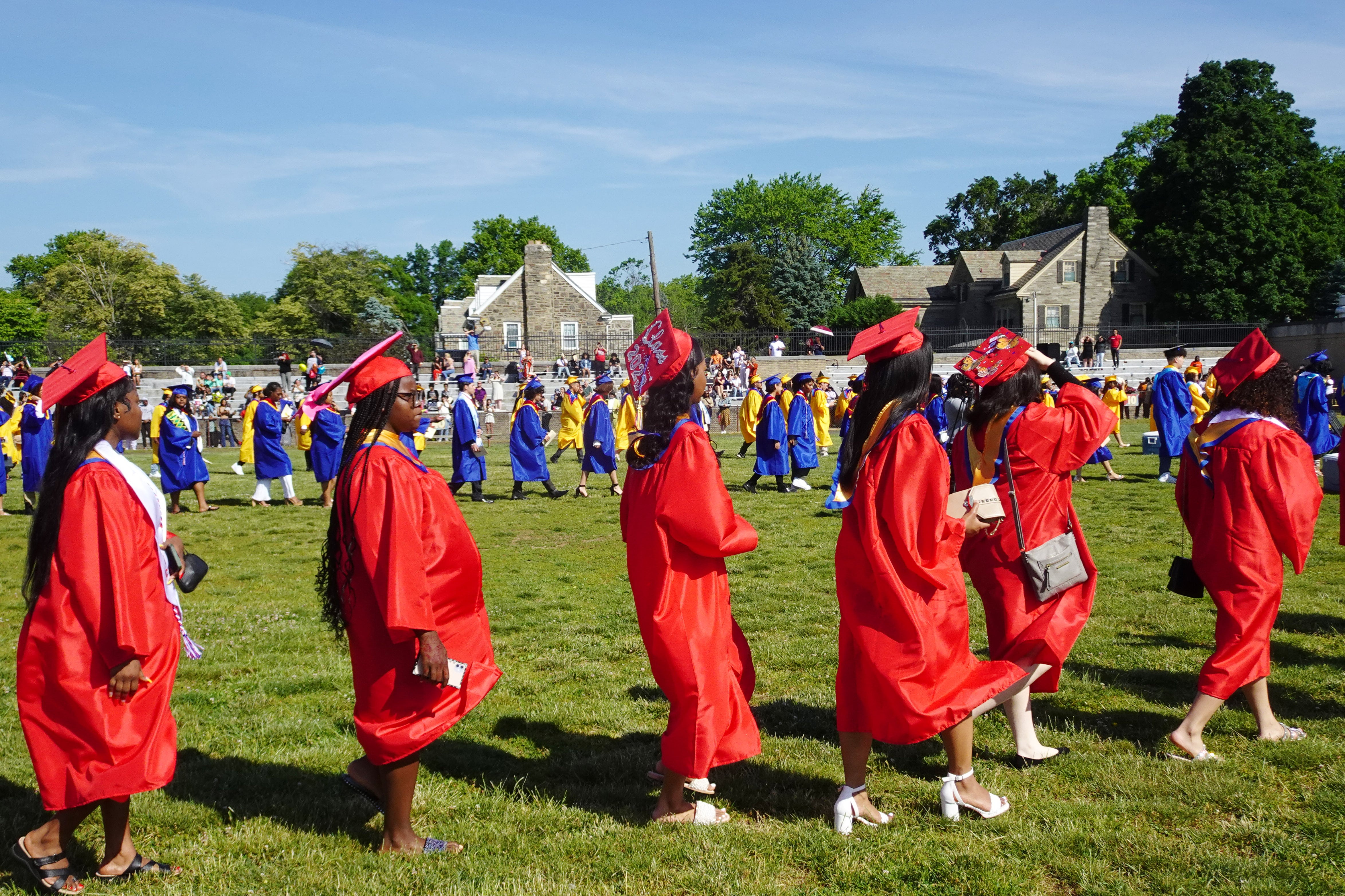 High school graduates in red gowns line up outside on a green grassy field with blue sky in the background.