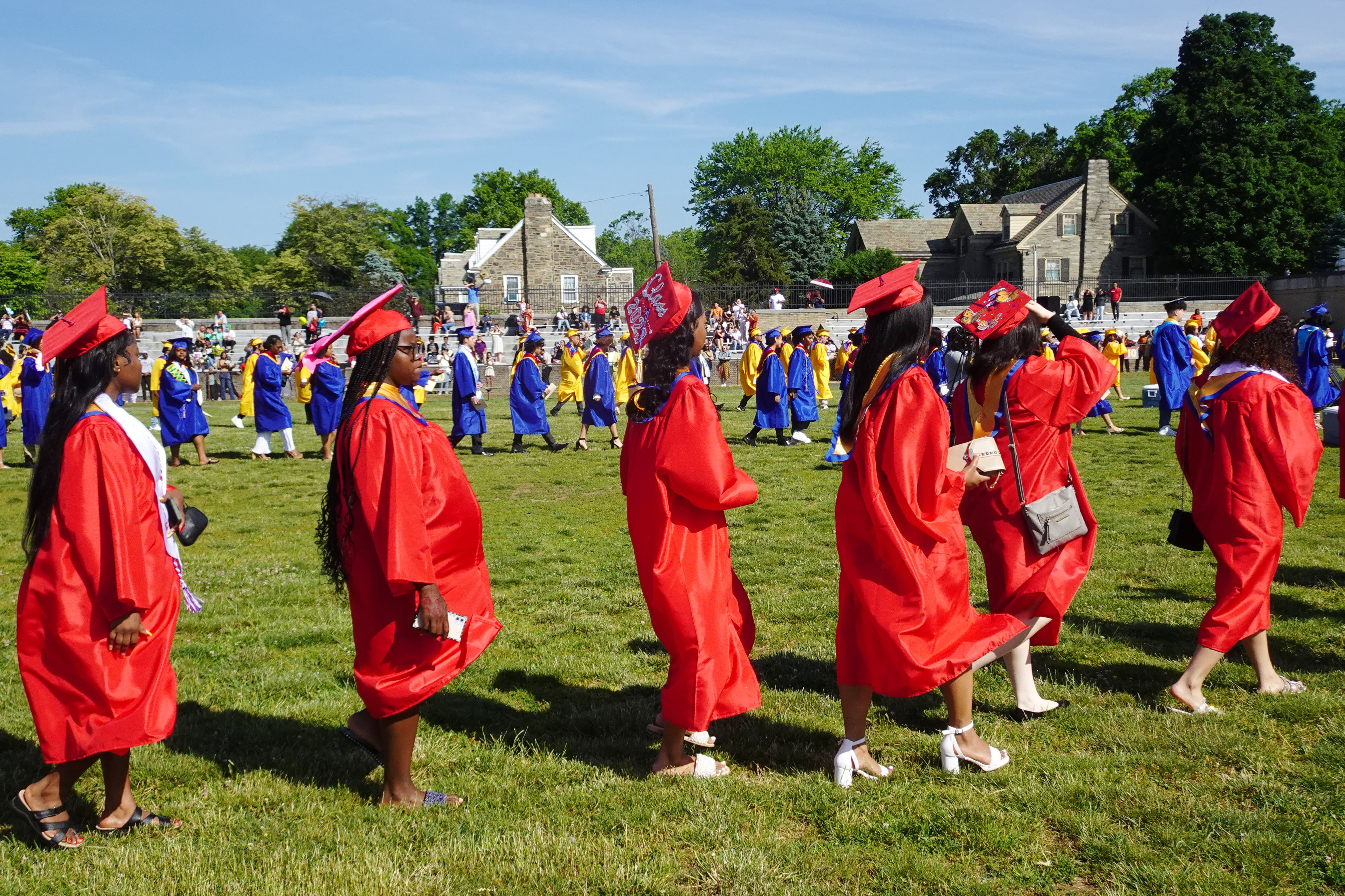 High school graduates in red gowns line up outside on a green grassy field with blue sky in the background.