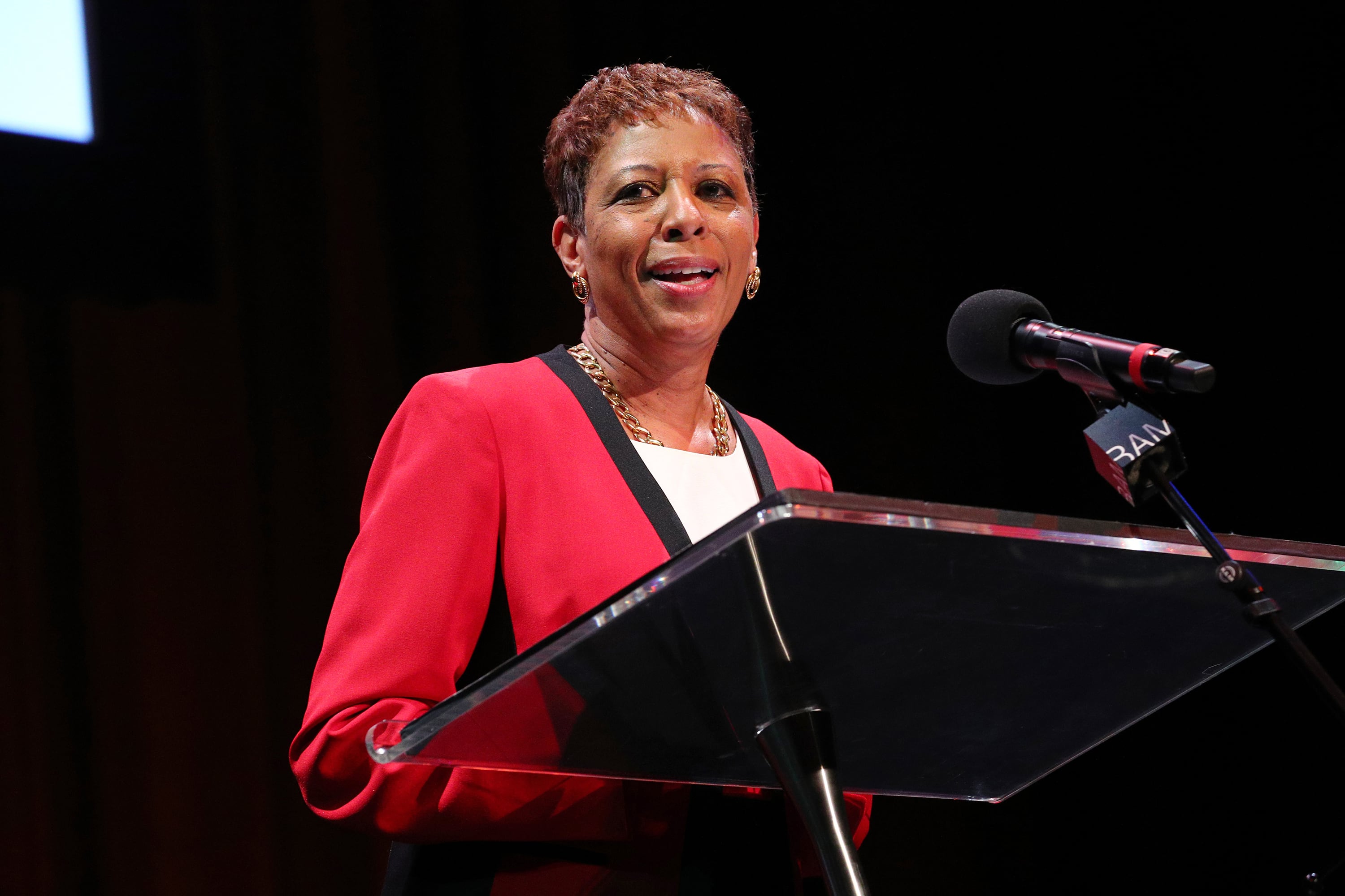 A woman with short dark hair and wearing a red suit jacket speaks behind a podium and into a microphone with a black background.