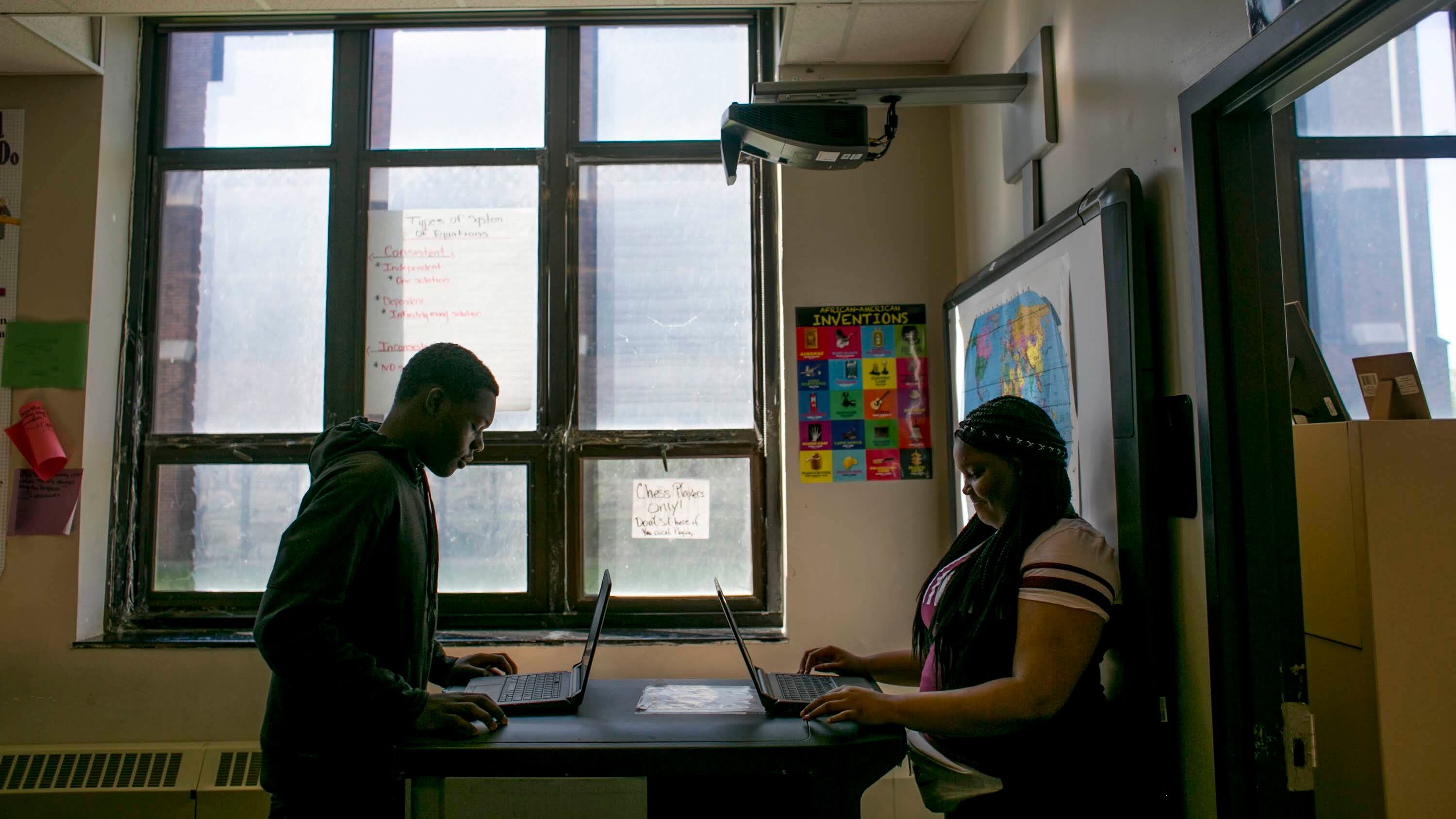 Two people sit in a shadowy classroom working on their laptops.