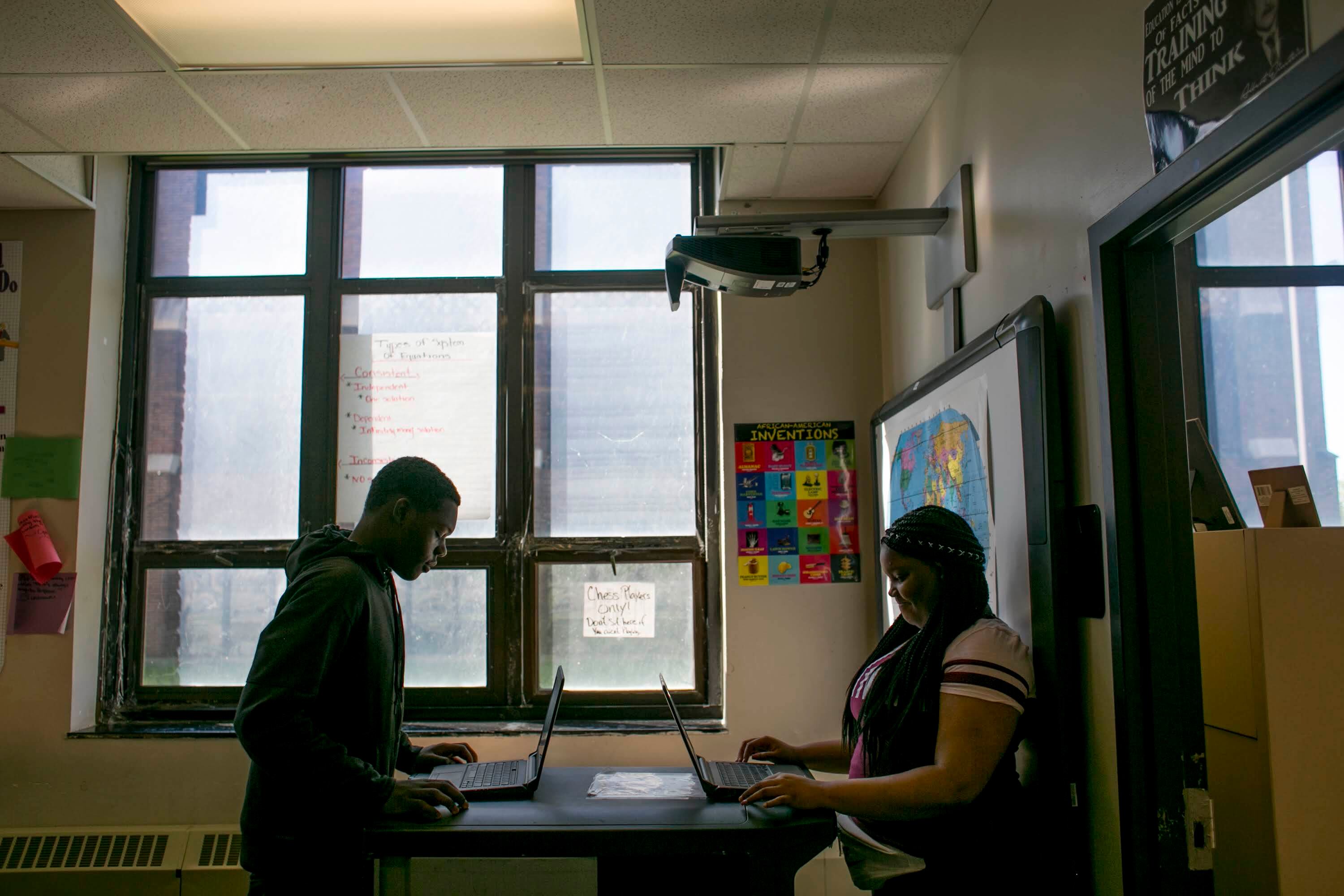 Two people sit in a shadowy classroom working on their laptops.