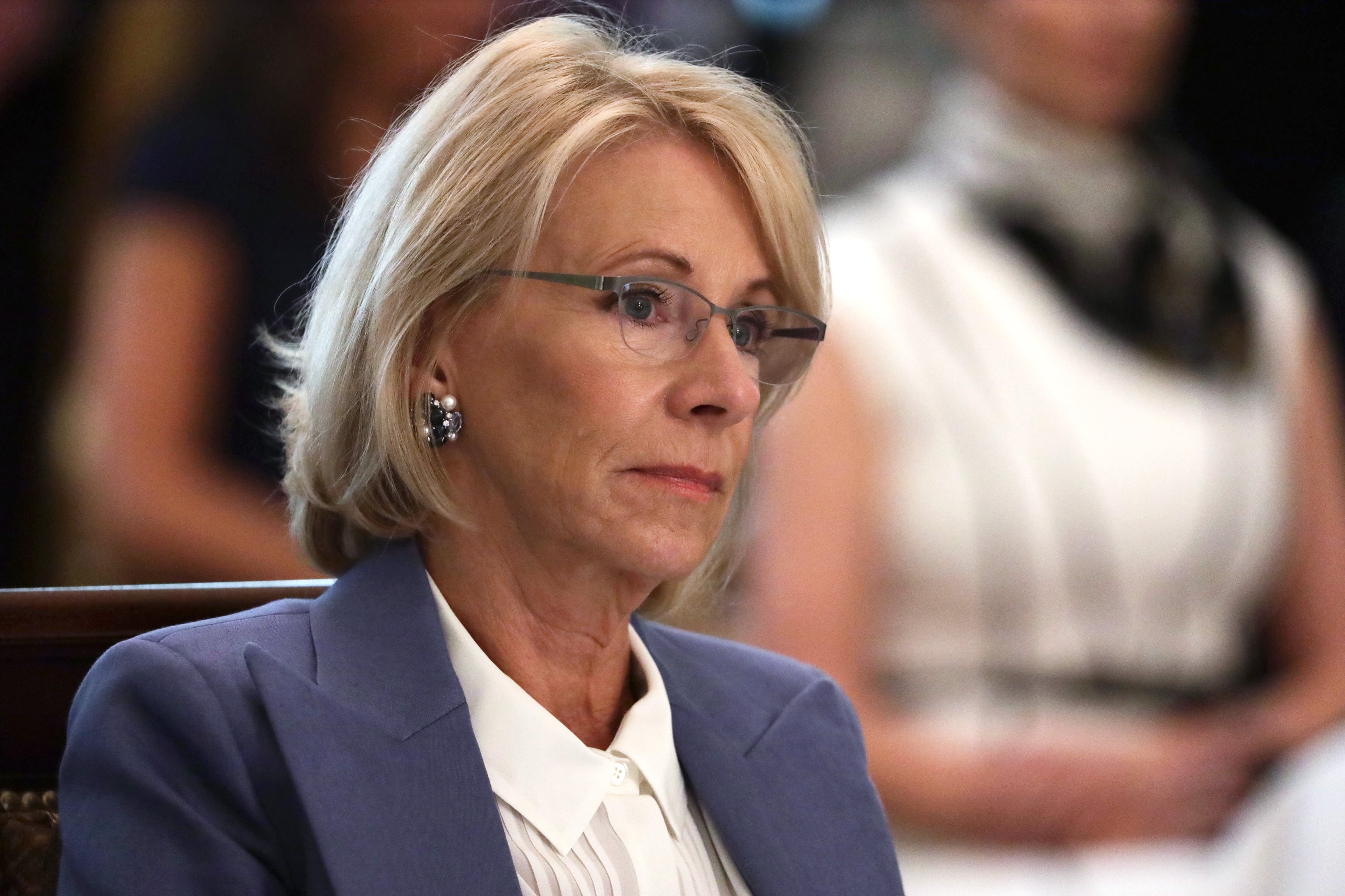 Betsy DeVos stares off in front of her, wearing glasses and a blue jacket.