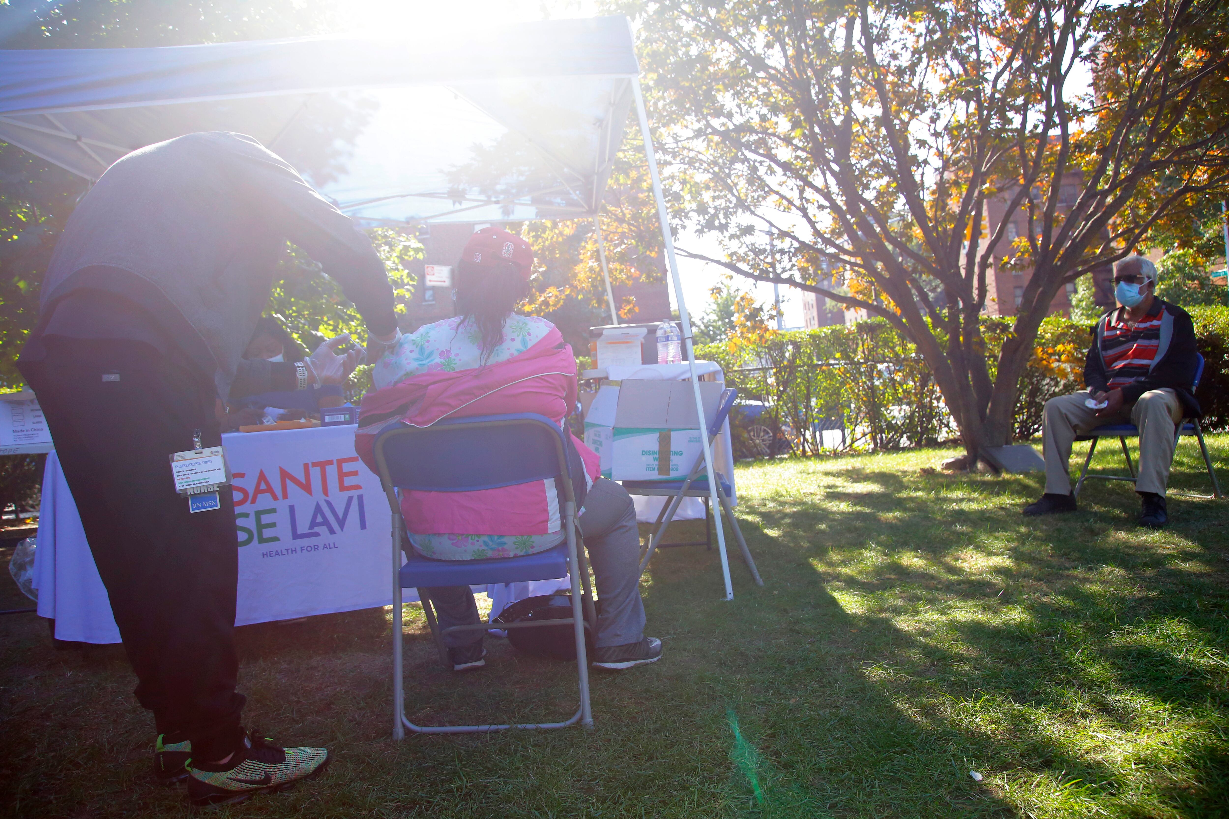 A medical professional gives a dose of the vaccine to a patient at an outdoor clinic as a man sits next to a tree behind them.