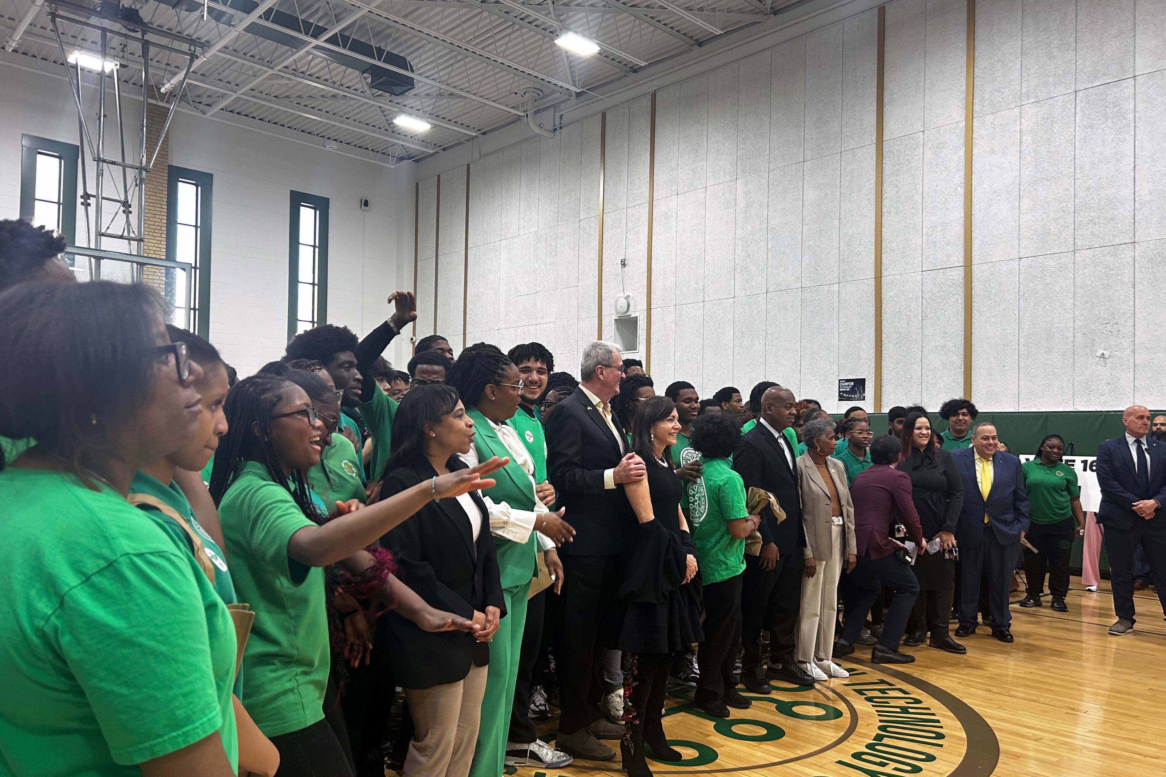 A group of people in suits and green t-shirts stand in a crowd in a school gym.