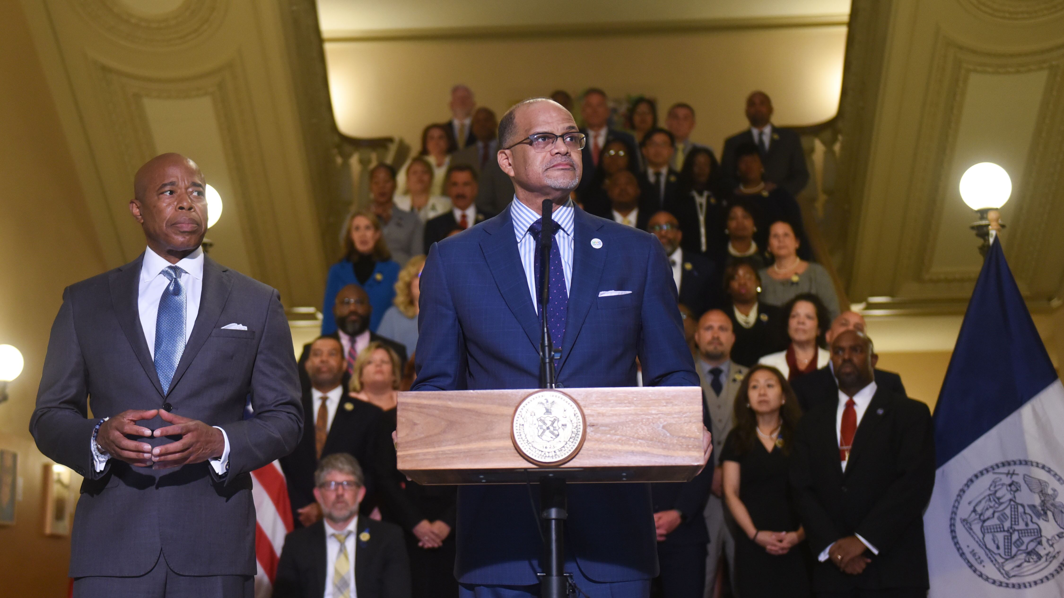 NYC Chancellor David Banks stands at a podium, with Mayor Eric Adams standing off to the left behind him.