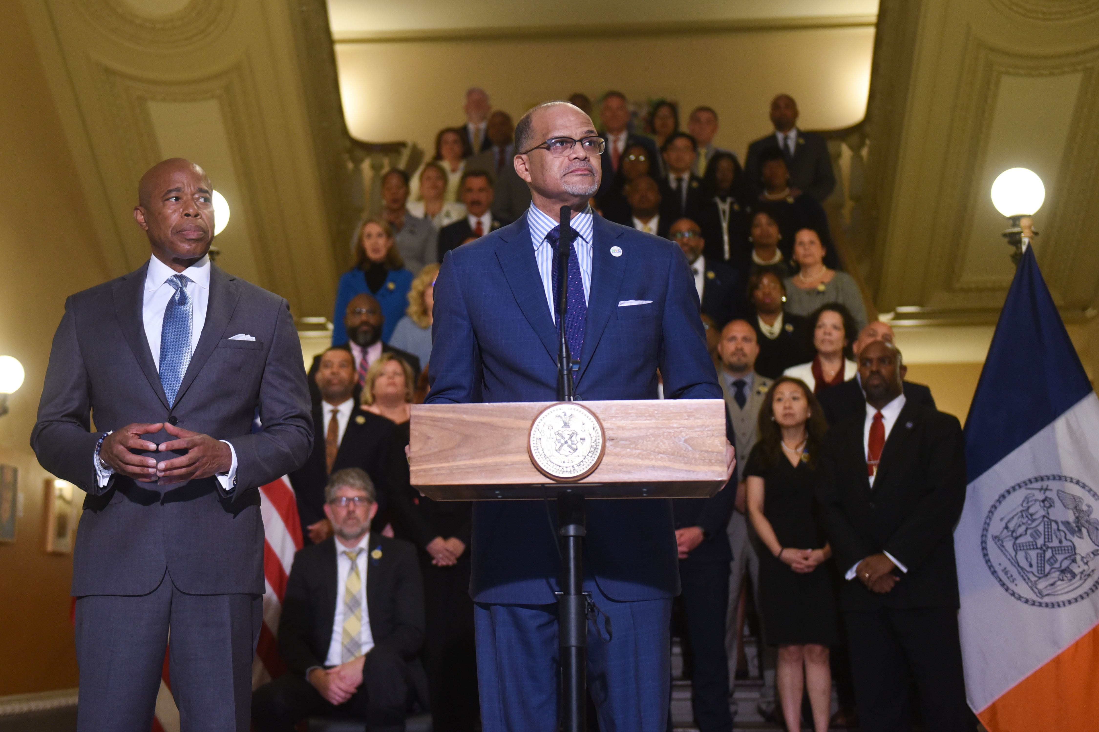 NYC Chancellor David Banks stands at a podium, with Mayor Eric Adams standing off to the left behind him.