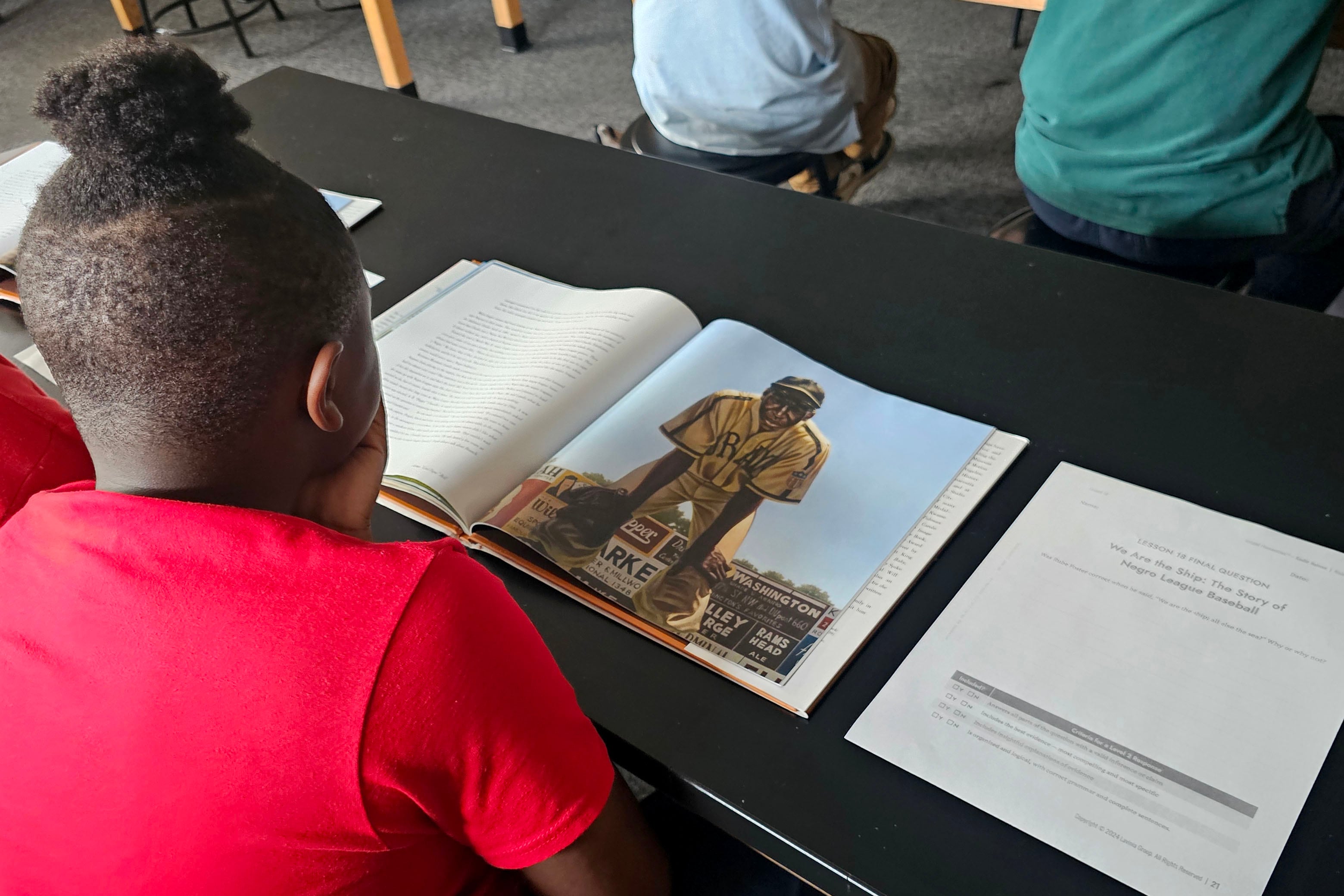 A student wearing a red shirt sits at a desk with an open book and worksheet on the top.