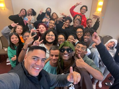 A large group of high school students and an adult pose for a selfie in a room.