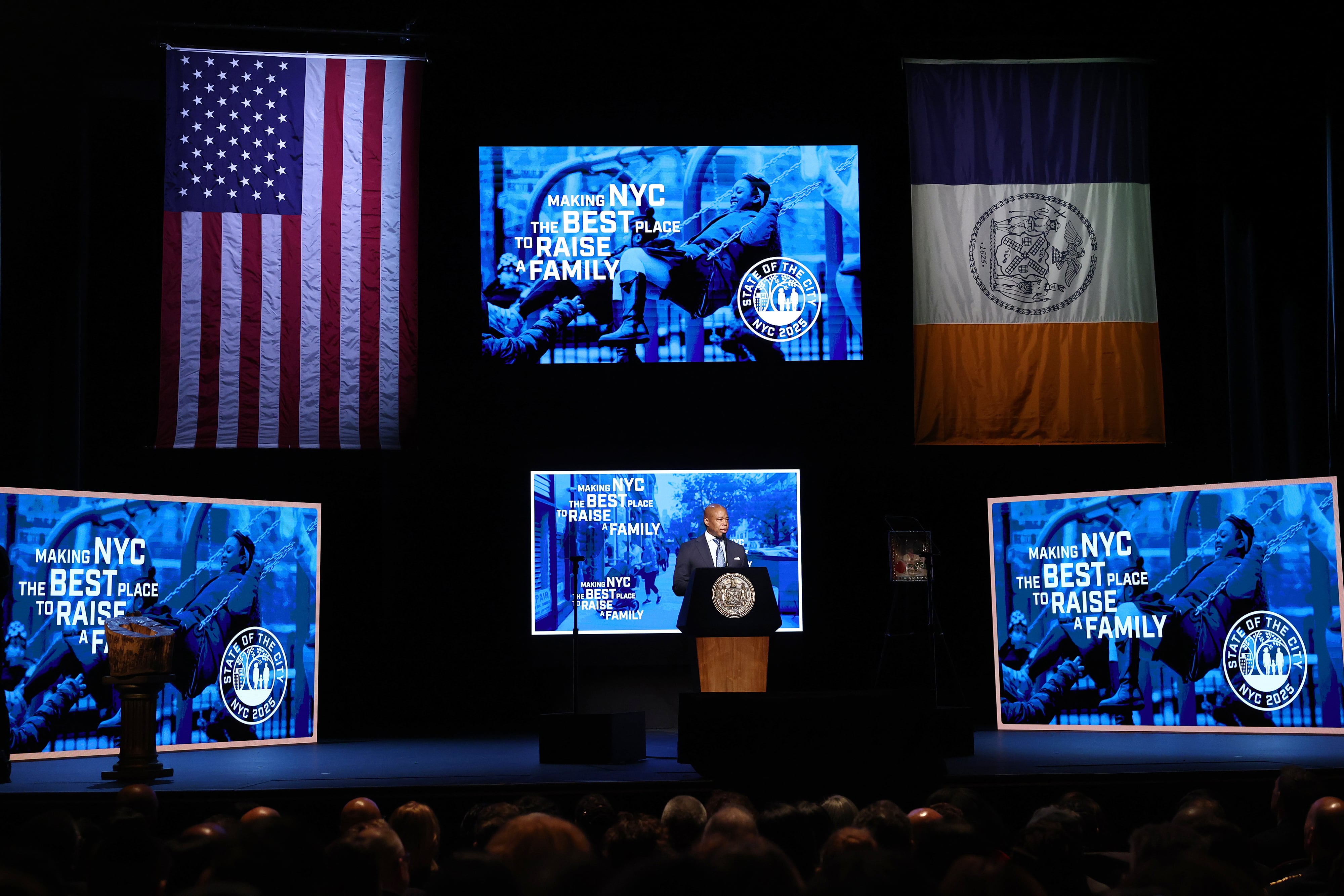 A man in a suit speaks from behind a podium in front of a large crowd with two large flags and three large screens in the background.