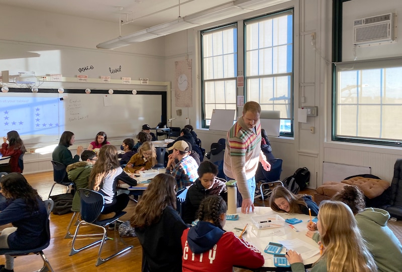 An adult teacher stands while a large group of students sit at their tables in a classroom with white walls in the background.
