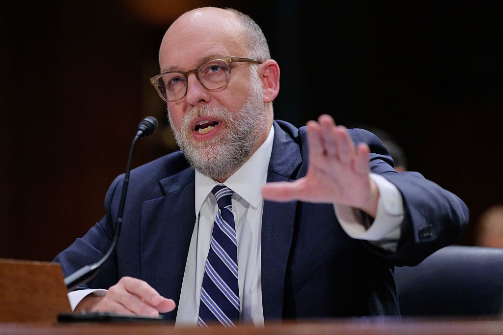 OMB Director Russ Vought, balding with close-cropped hair, glasses and a salt and a pepper beard, sits at a desk and speaks into a microphone. He's wearing a dark blue suit and striped tie. He's gesturing with his left hand.