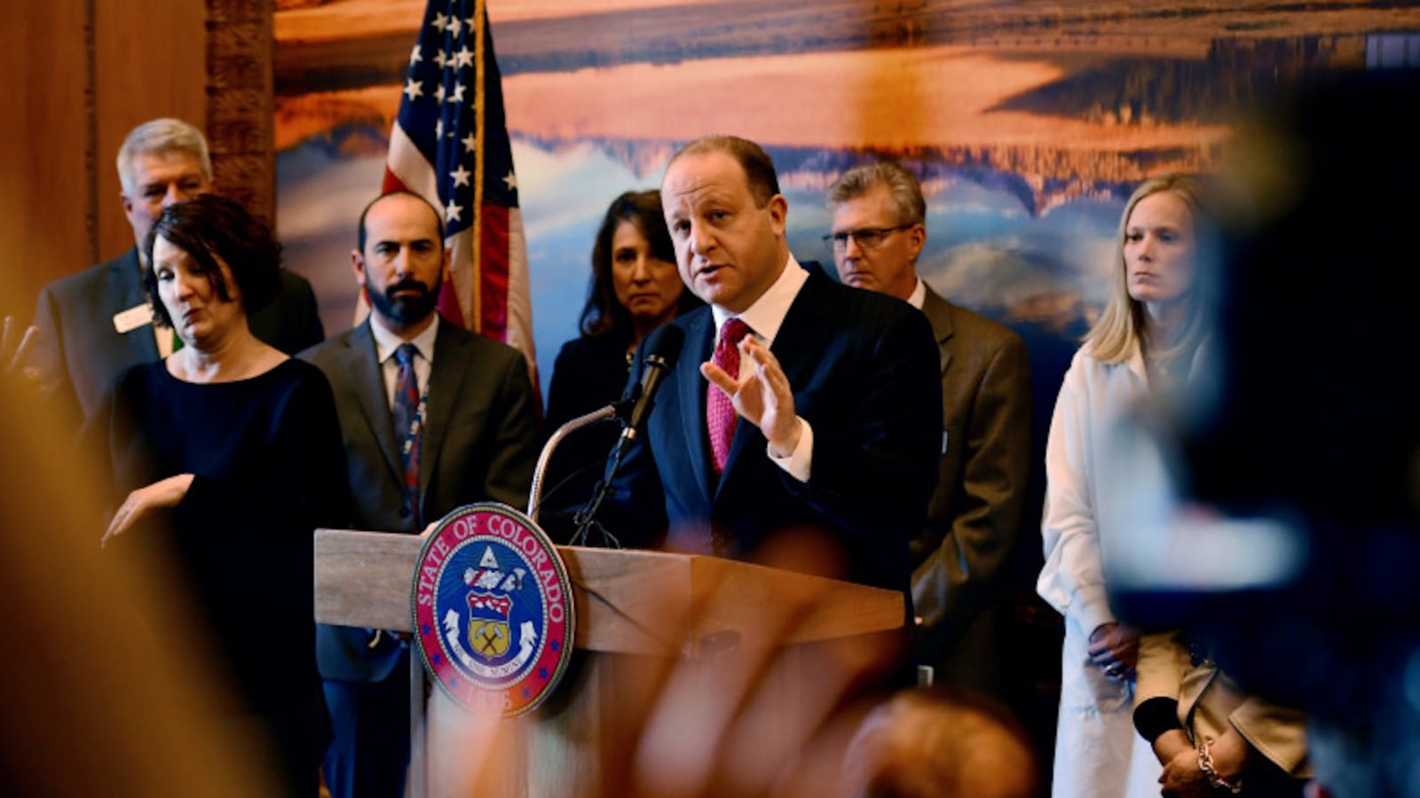 Colorado Governor Jared Polis speaks at a State of Colorado podium, surrounded by several people.