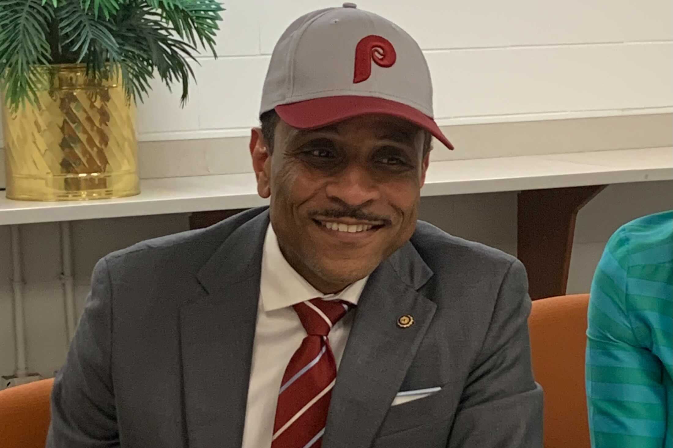 A man in a red and white tie and gray suit is wearing a Philadelphia Phillies hat while seated at a table.