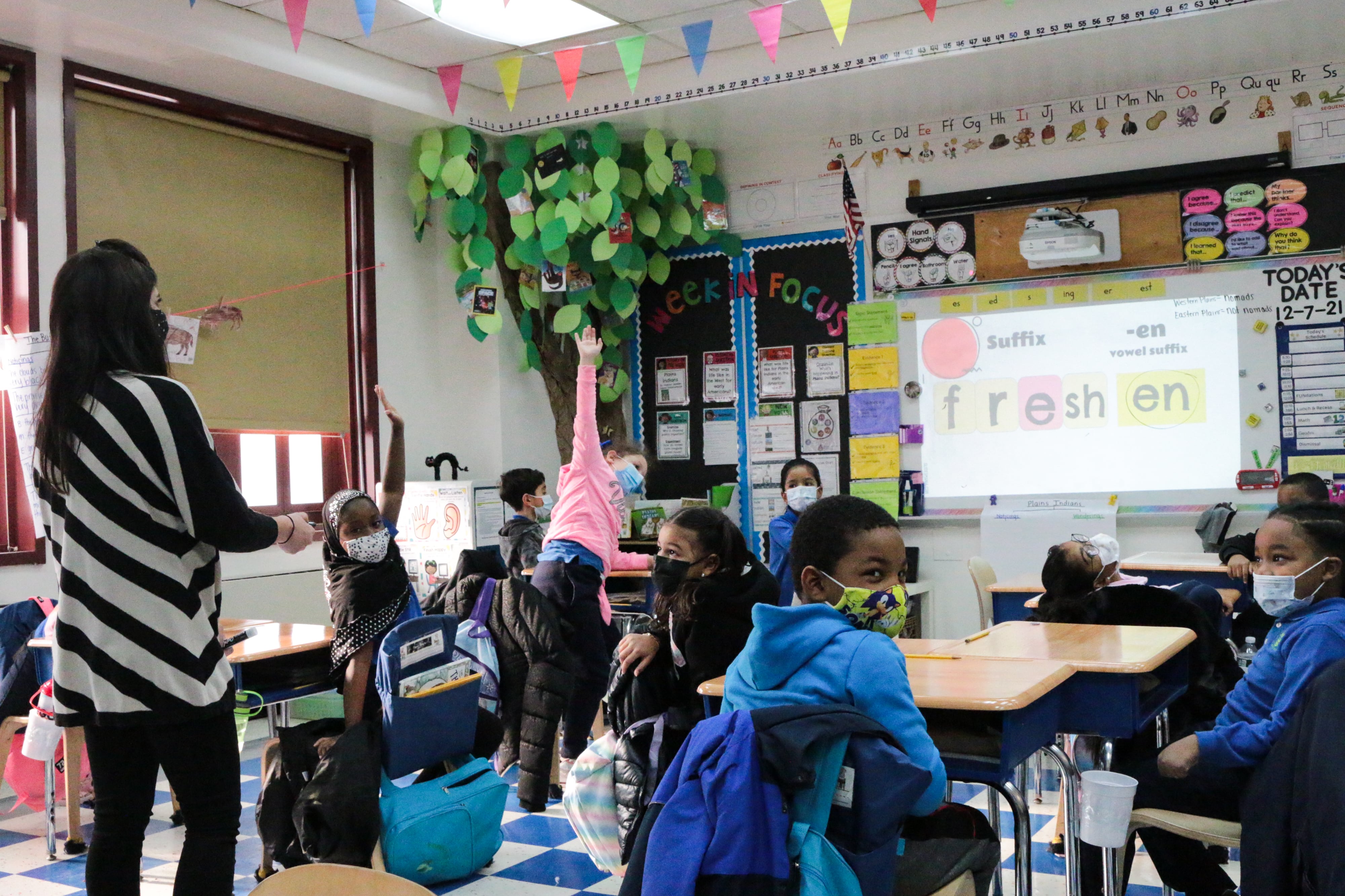 A teacher works with students on a reading exercise; students emphatically raise their hands as the projector on a white board reads “freshen.”