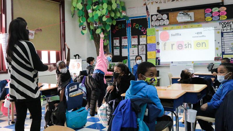 A teacher works with students on a reading exercise; students emphatically raise their hands as the projector on a white board reads “freshen.”