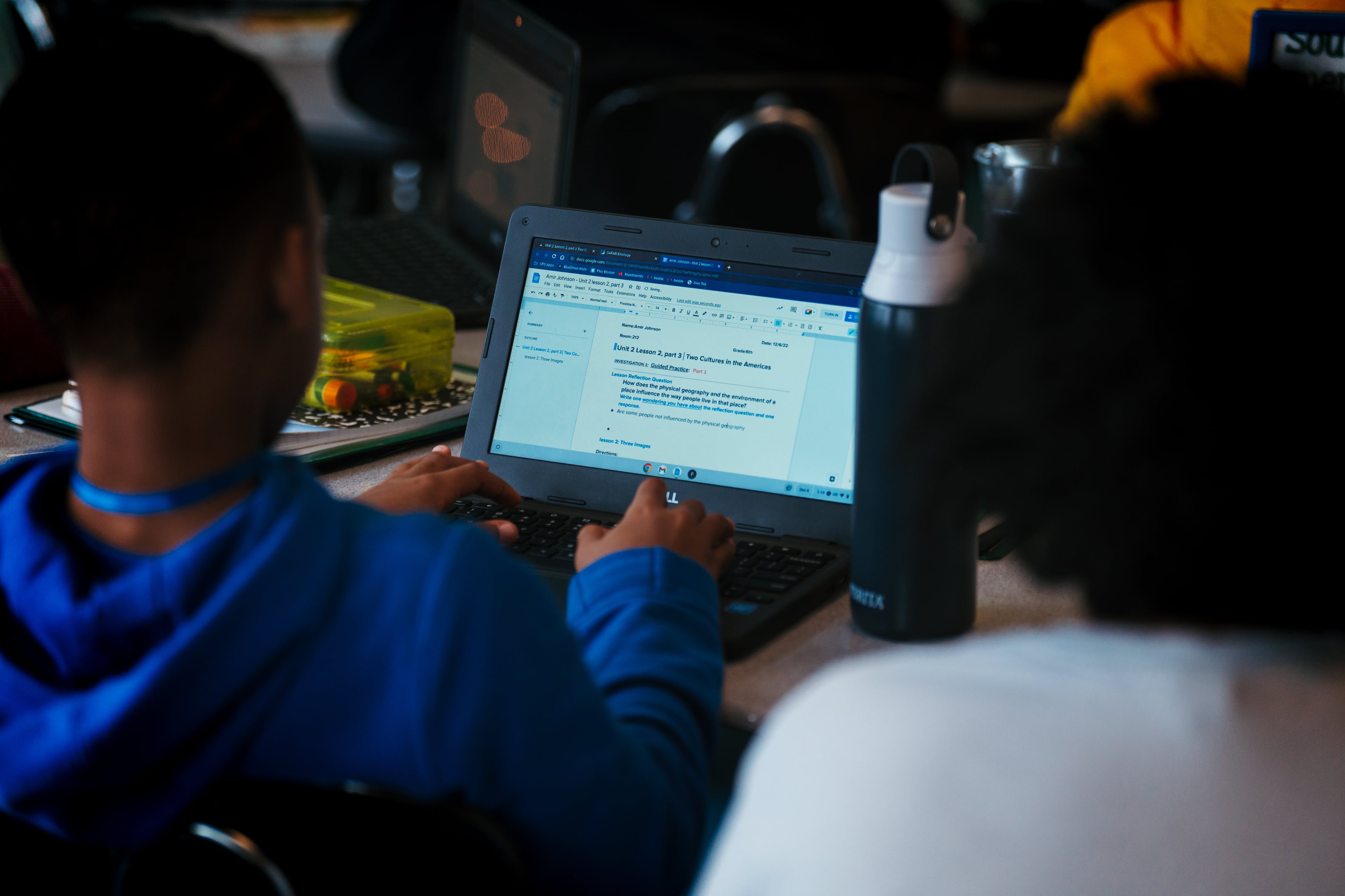 A photograph of the back of a middle school student wearing a blue sweater typing and reading from a laptop at a desk in a classroom.