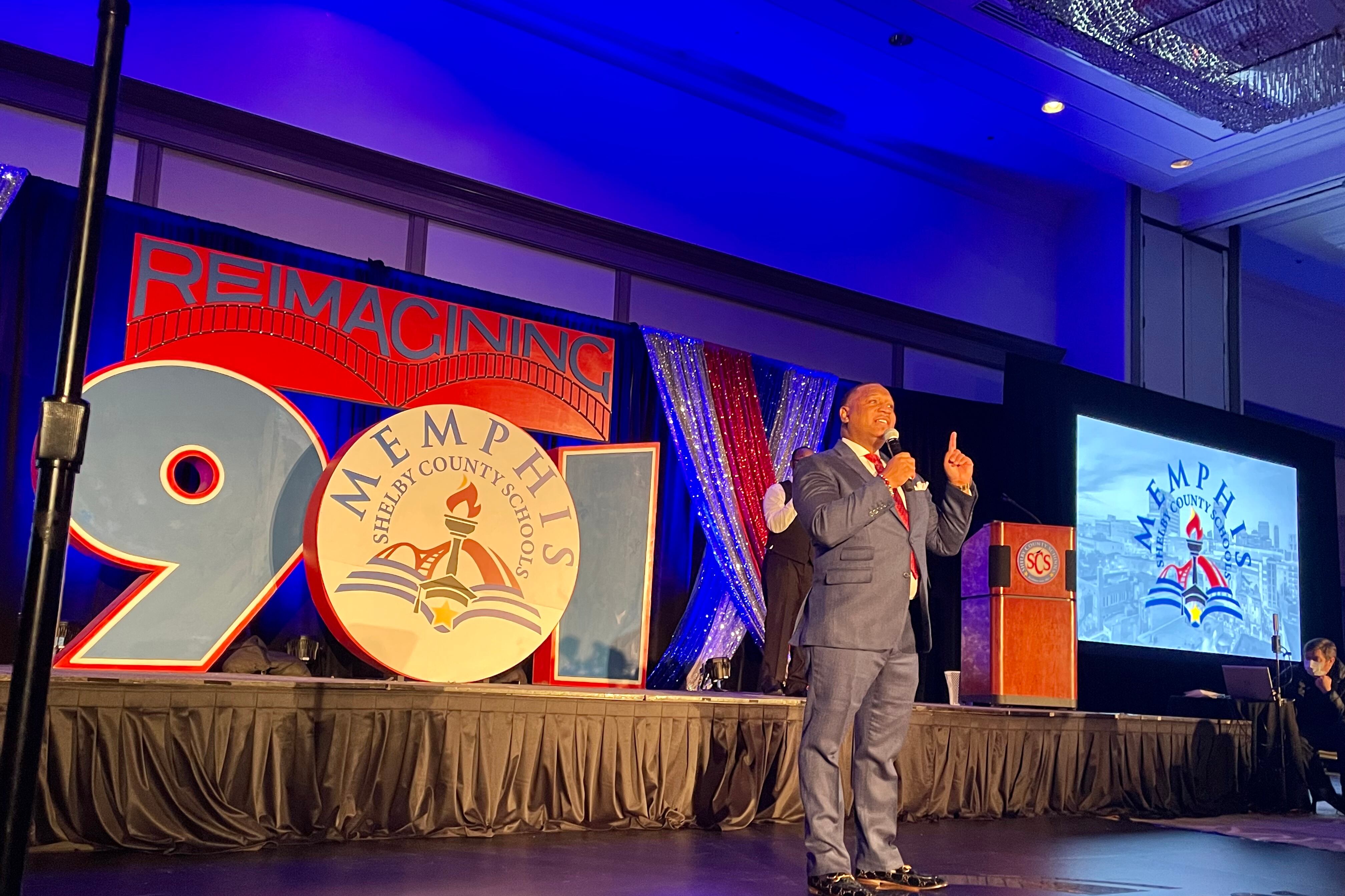 A man wearing a suit points up as he speaks into a microphone