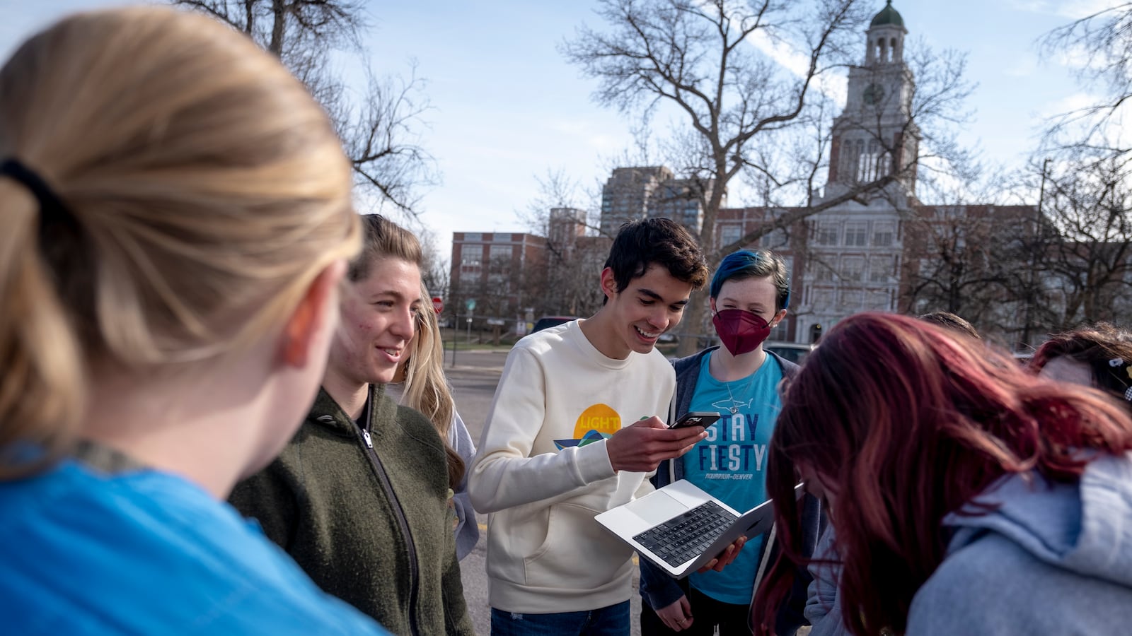 A group of high school students laugh as they plan an event, standing in the parking lot of their high school.