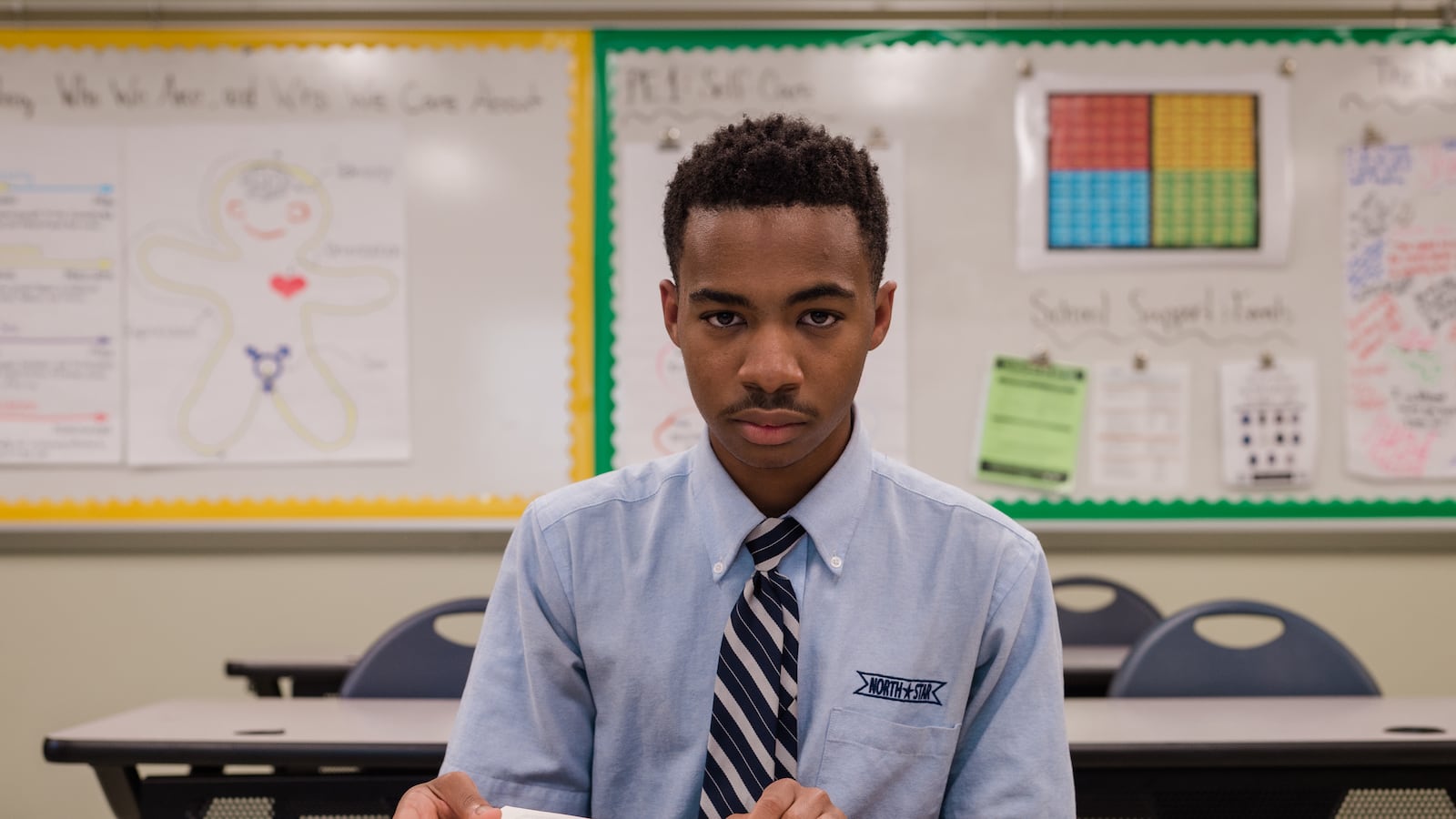 Photo of a teenage boy wearing a blue button-down shirt and a tie. He sits at a desk in a classroom. 