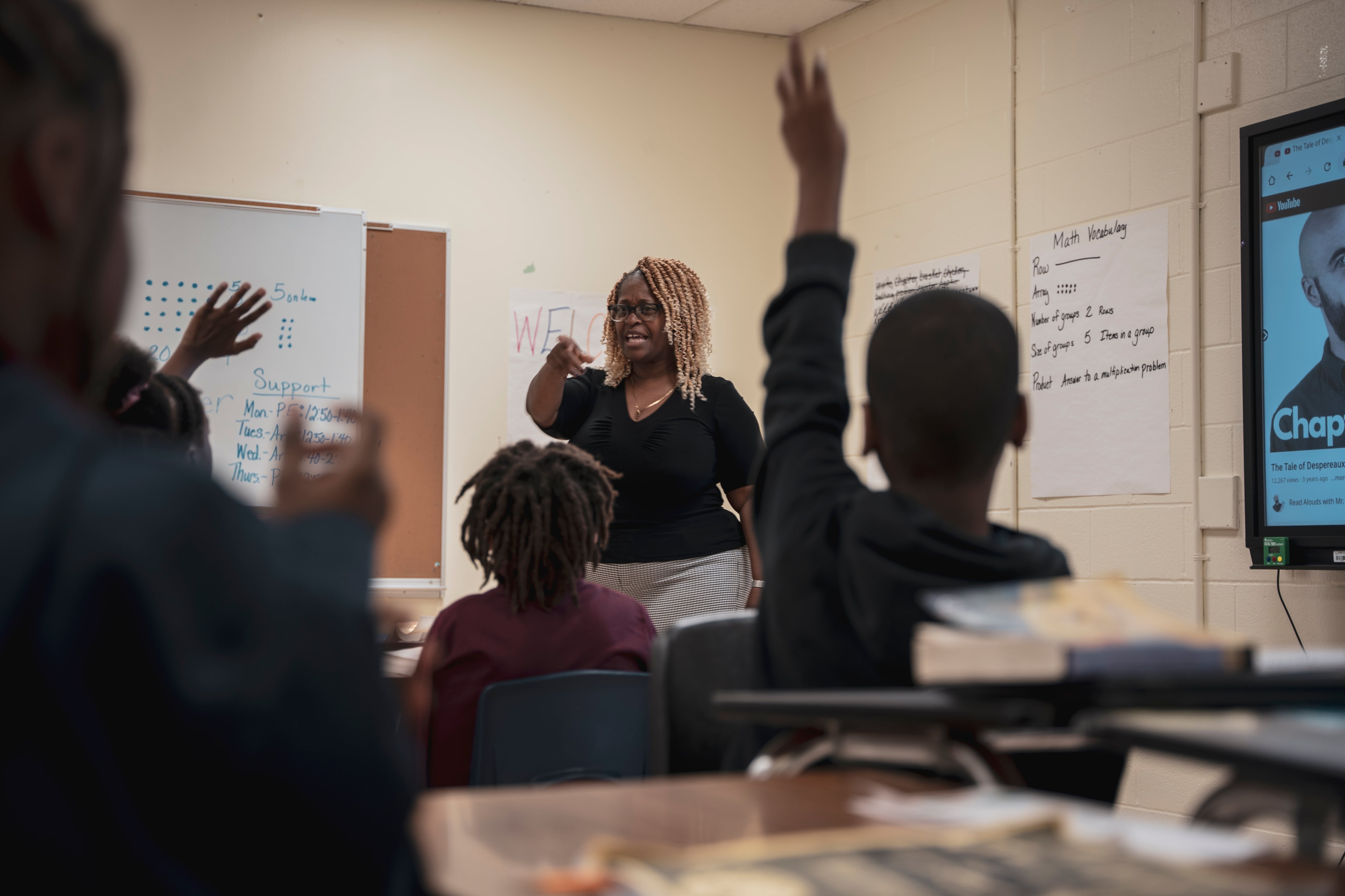 A woman stands at the front of a classroom with a few students in the foreground of the frame.